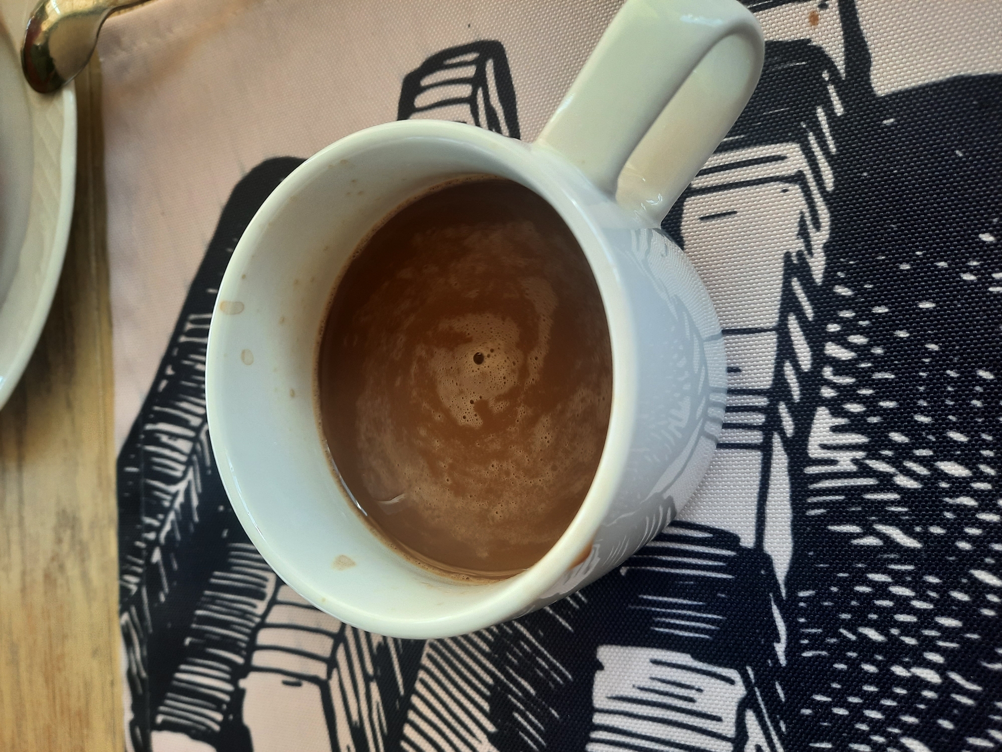 A white mug filled with hot chocolate sits on a table. The mug has a handle and a textured pattern. The hot chocolate has a foamy surface with small bubbles. The mug is placed on a patterned placemat with black and white abstract designs. In the background, there is a wooden surface and a white plate with a silver spoon. The overall scene suggests a cozy and relaxed setting for a warm beverage. The image is set in an unspecified location in Ukraine.