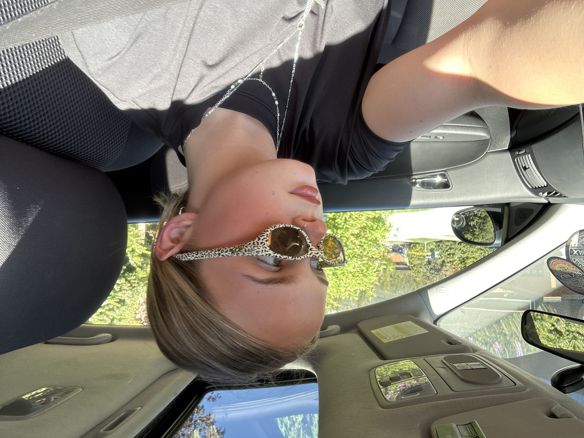 A woman with light brown hair and sunglasses is sitting in the driver's seat of a car. The car is parked outside and there are trees visible through the windshield. The woman is wearing a black shirt and a silver necklace. The interior of the car is gray. The location cannot be determined to be Whistler, Canada from the image provided.