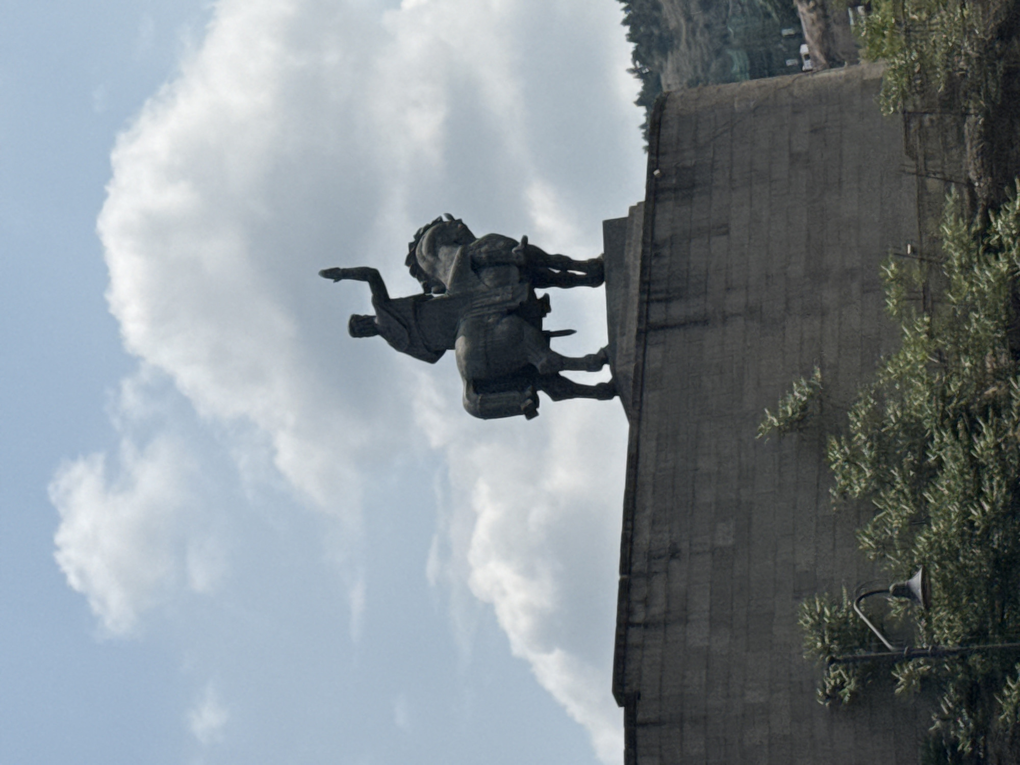 The image depicts a monumental equestrian statue of Vakhtang Gorgasali, the founder of Tbilisi, Georgia, set against a backdrop of a cloudy blue sky and a distant hillside with trees. The statue, cast in a dark material, shows Gorgasali seated on a horse, with his right arm raised. The statue is positioned atop a large, grey stone wall, which forms the foreground of the image. A vintage-style lamppost with a curved arm and a dark lampshade stands to the left of the wall, partially obscured by lush green foliage. The wall itself appears to be constructed from large, rectangular blocks of stone. The overall scene suggests an outdoor public space in Tbilisi, likely a historical monument or landmark.
