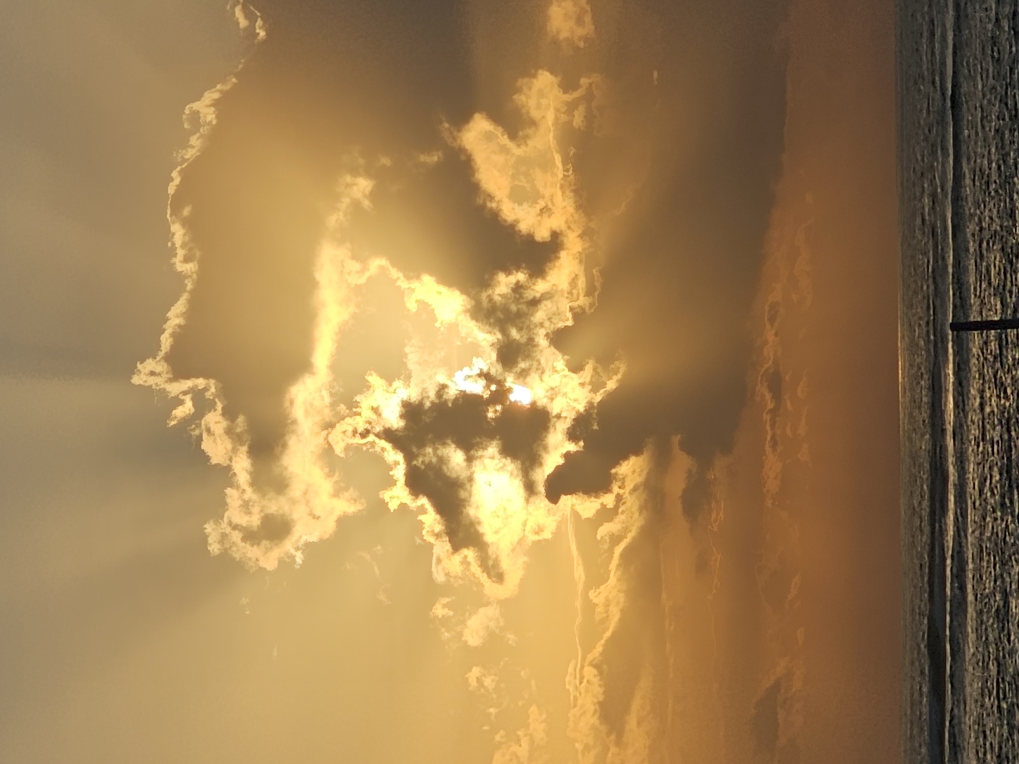 A serene sunset scene unfolds over the ocean near Denpasar, Indonesia. The sky is awash in warm, golden hues as the sun, partially obscured by dramatic clouds, casts its light through them in visible rays. The ocean below reflects the ambient light, with gentle waves creating a textured surface. The horizon line is clearly defined, separating the vast expanse of the sky from the tranquil sea. No people or specific man-made structures are discernable in this natural landscape, suggesting a peaceful and unpopulated coastal view.