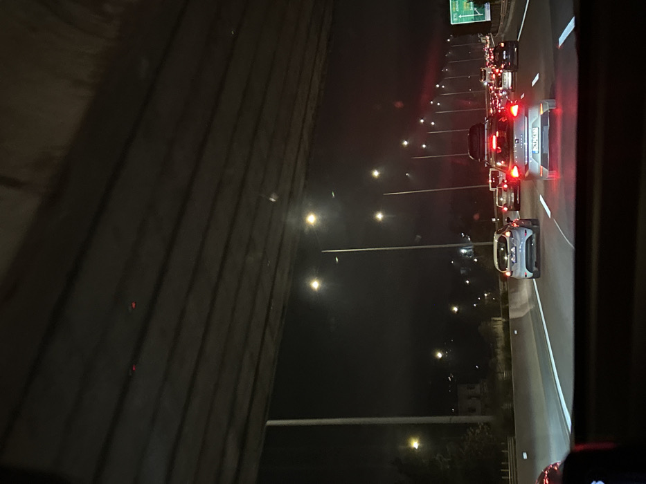 The image captures a night scene on a multi-lane highway, viewed from inside a vehicle. The perspective is rotated, showing a dark, textured wall or barrier on the left side, running vertically along the frame.
On the right side of the image, a busy highway stretches into the distance, filled with vehicles. Numerous red taillights from cars in traffic are visible, indicating either slow-moving or stopped conditions. Bright streetlights line the highway, casting illumination on the road and surrounding area.
In the foreground, a light-colored car (possibly white or silver) is prominent, with its rear facing the viewer. It appears to be equipped with a roof rack. Another car is visible directly behind it, and a long line of additional vehicles extends further down the road.
A green highway sign, partially obscured and indistinct, can be seen in the upper right portion of the image, suggesting an upcoming exit or destination. The dark sky confirms it is nighttime. The weather appears clear, with no visible precipitation or fog. No people are distinctly visible, and any text on the distant highway sign or vehicle license plates is illegible due to distance and image quality. The overall impression is one of night-time traffic on a major road.