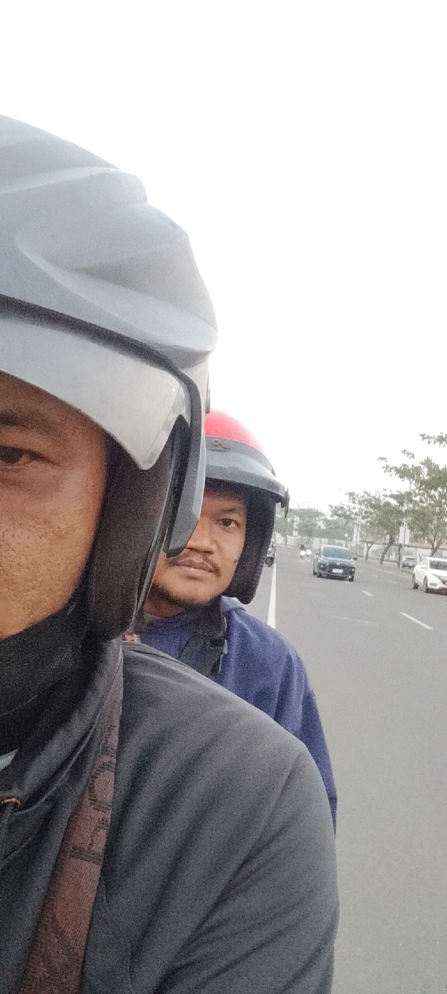 Two individuals are seen from the chest up, both wearing motorcycle helmets, in what appears to be a selfie. The person in the foreground, on the left, has a light grey helmet and is wearing a dark grey hooded jacket with a patterned strap visible across the chest. Their face is partially visible on the left side of the frame. The second person, in the background, is wearing a red and black helmet and a blue hooded sweatshirt. Their face is clearly visible, looking towards the camera. They have a mustache and a short beard. The background suggests an urban environment with a road, several cars, and trees lining the side. The sky is overcast and appears pale. The scene is consistent with a view from a moving motorcycle on a road in Gunung Putri, Indonesia.