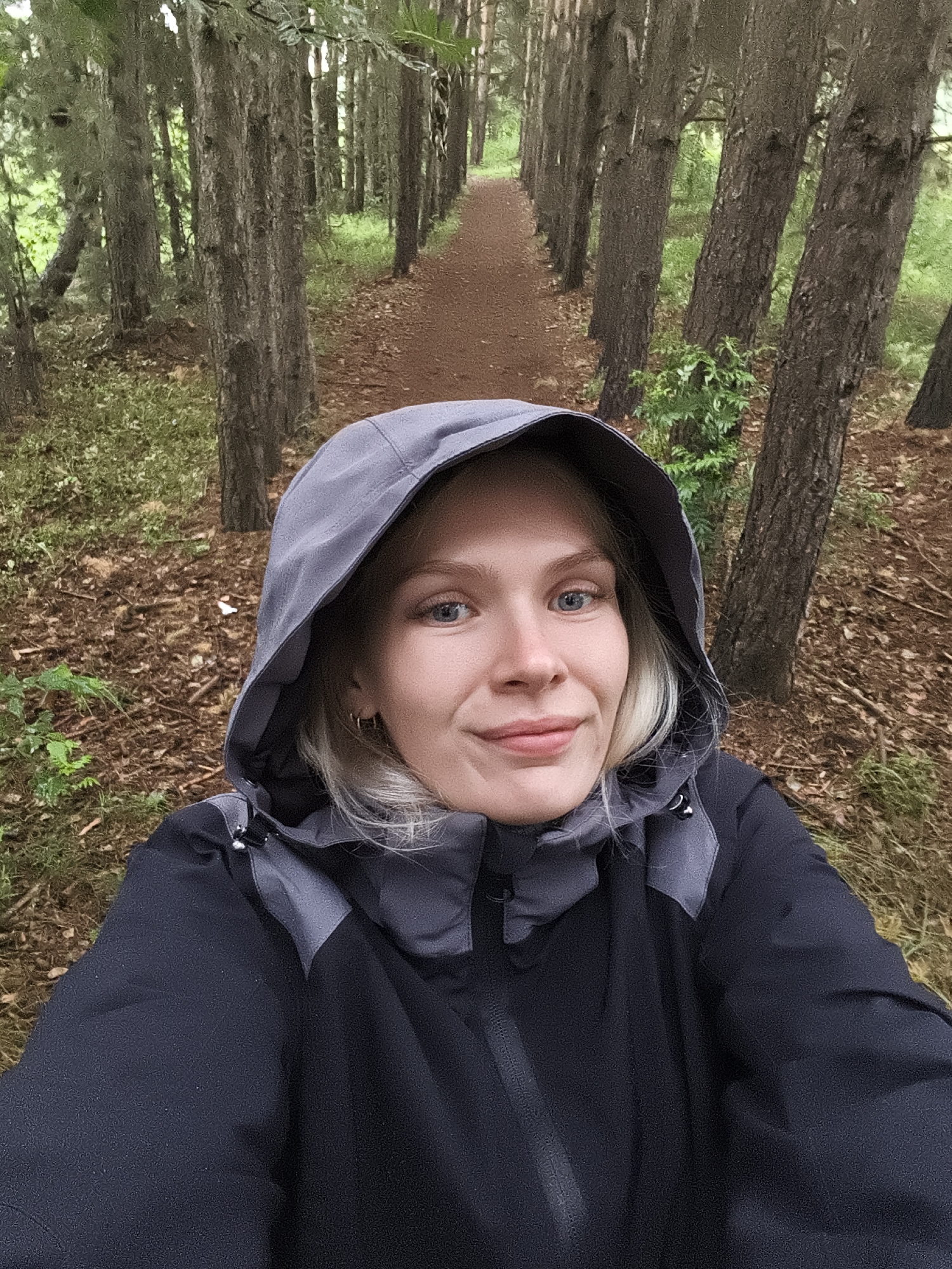 In Русский Вожой, Russia, a young Caucasian woman with blonde hair and blue eyes is taking a selfie outdoors in a forest. She is wearing a dark grey hooded jacket with grey accents. The background features a pathway lined with tall, slender trees, with fallen leaves and some green undergrowth on the forest floor. The lighting suggests an overcast day.