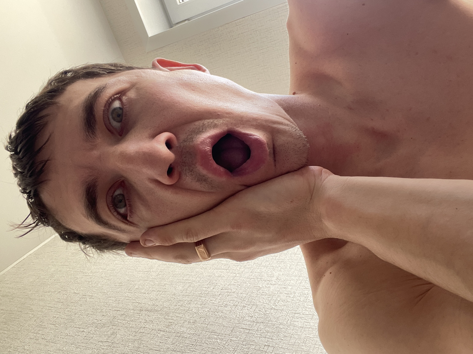 A close-up, overhead, selfie-style shot shows a Caucasian man with brown hair and open mouth. The man is shirtless, with his right hand on his left cheek. He is wearing a gold ring on his right ring finger. The background appears to be a textured, light-colored wall. The lighting is bright, suggesting it is daytime. There is no indication of the location in the city of Laishevo, Russia.