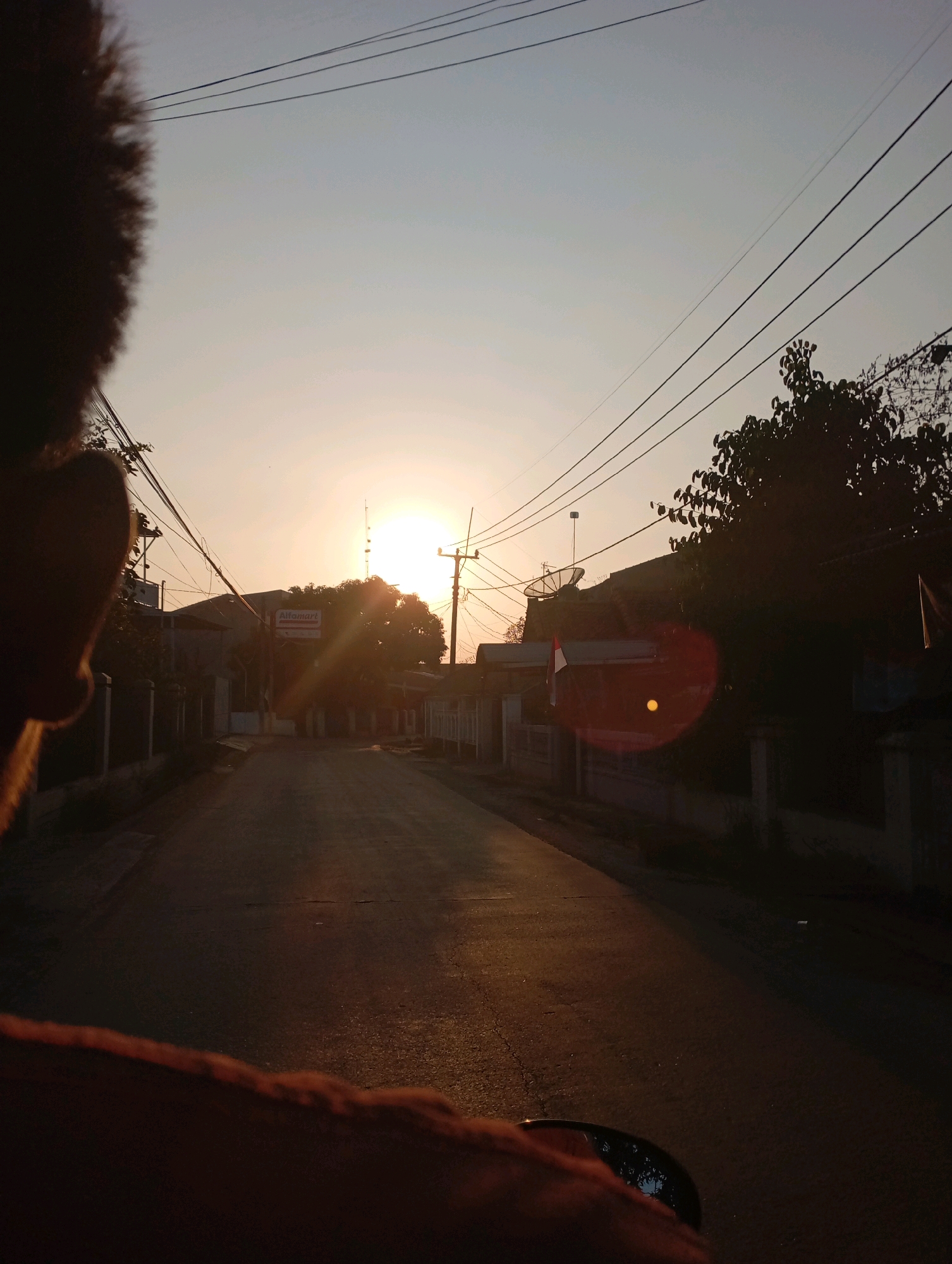 A side-view, first-person perspective shot taken from a motorcycle, captures a road in Kecamatan Haurgeulis, Indonesia, during sunset. The sun is low in the sky, casting a warm, golden light across the asphalt road and illuminating the surrounding residential area. Overhead, multiple electrical wires crisscross the frame, supported by poles. On the right side of the road, a large tree partially obscures the view of houses and a satellite dish mounted on a rooftop. A circular lens flare is visible in the mid-right portion of the image. In the background, a sign for "Alfamart" is visible on a building. The foreground shows the edge of a person's face and shoulder on the left, and the side of the motorcycle's mirror and a hand on the right. The overall scene depicts a common streetscape in a Southeast Asian town at dusk.