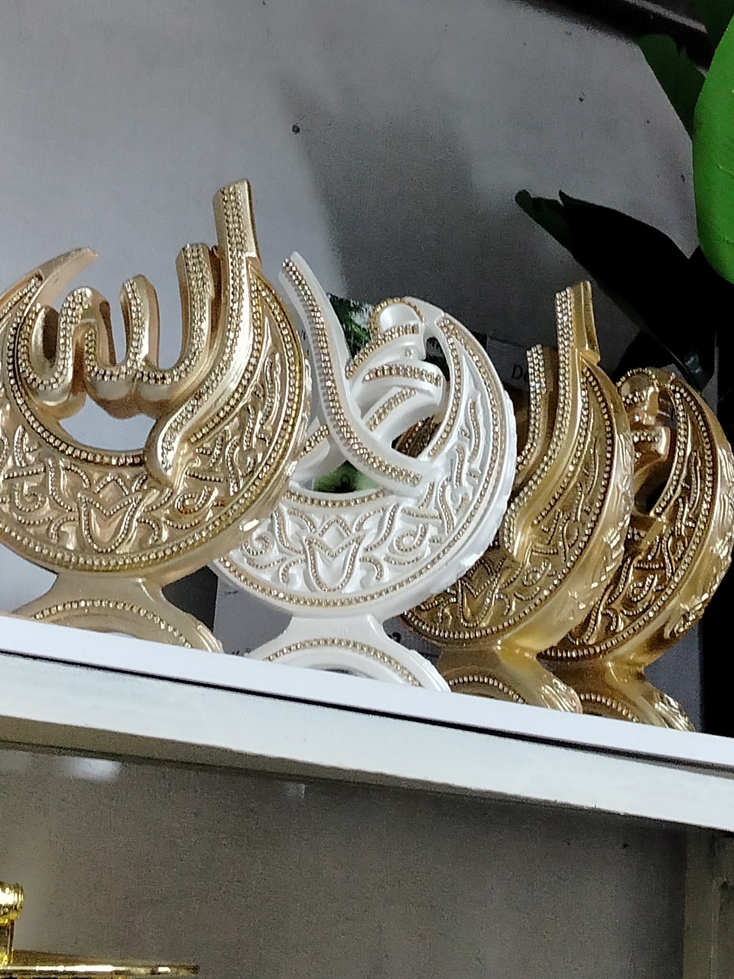 The image displays a shelf featuring decorative Islamic calligraphy ornaments. In the foreground, there are three such ornaments. The leftmost ornament is gold and appears to be inscribed with Arabic script. To its right is a white ornament, also with Arabic script and decorative detailing. The rightmost ornament is gold and features similar Arabic script and intricate patterns. A fourth, partially visible gold ornament is positioned behind the white one and to the right. The background shows a wall and a hint of a green leaf from a plant. The shelf is white and positioned against a gray wall. The location is identified as Көлкент, Kazakhstan.