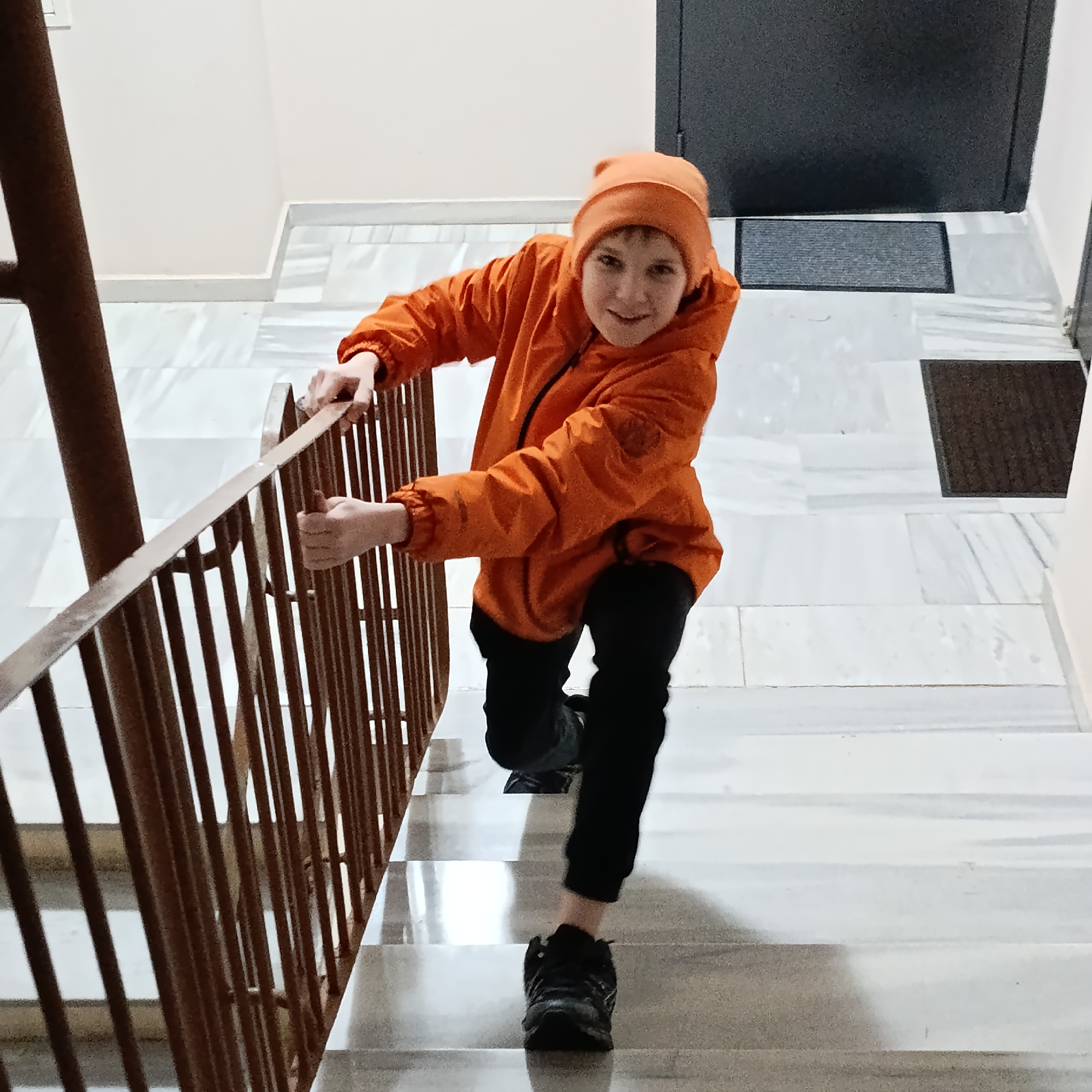 A boy wearing an orange hooded jacket and black pants climbs a set of stairs. He is holding onto the metal railing on his left. The stairs are made of white marble with grey veining. Behind him is a closed, dark-colored door with a grey doormat in front of it. The environment is an indoor stairwell. It is unknown if the location is in the city of Марковский, Russia.