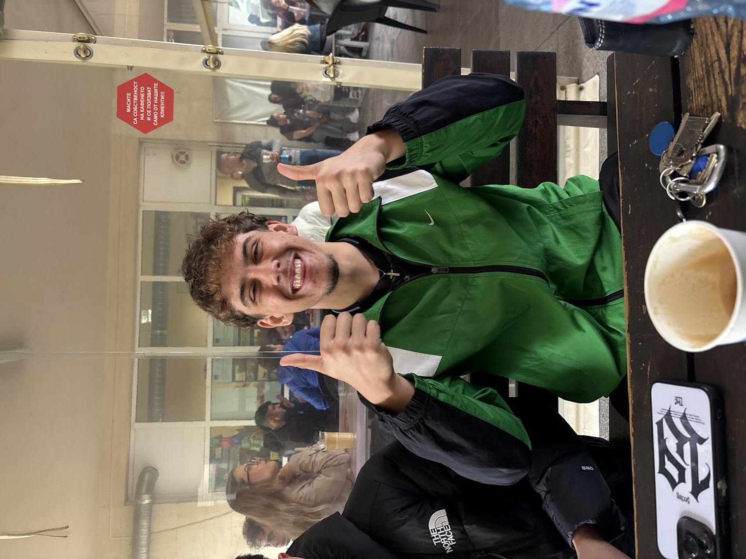 The image captures a young man seated at a dark wooden table indoors, likely in a cafe or restaurant in Plovdiv, Bulgaria. He has curly brown hair and a short beard, is smiling broadly, and gives a double thumbs-up to the camera. He is wearing a green, black, and white jacket with a Nike logo.
On the table next to him are a set of keys, a white cup containing a light-colored beverage (possibly coffee), and a smartphone in a white case featuring a black gothic-style design and the word "THE". A clear glass partition runs across the scene, reflecting parts of the room and other patrons in the background, who appear to be socializing or waiting.
A prominent red sign, visible through the glass, displays Bulgarian text: "МАСИТЕ СА СОБСТВЕНОСТ НА КАФЕНЕТО И СЕ ПОЛЗВАТ САМО ОТ НАШИТЕ КЛИЕНТИ!!!" This translates to "THE TABLES ARE PROPERTY OF THE CAFE AND ARE USED ONLY BY OUR CUSTOMERS!!!", clearly indicating the commercial nature of the establishment. Other visible text includes "NIKE" on the subject's jacket and "THE NORTH 550" (likely "The North Face") on another jacket reflected in the glass. The scene conveys a cheerful and casual atmosphere, possibly during the day.