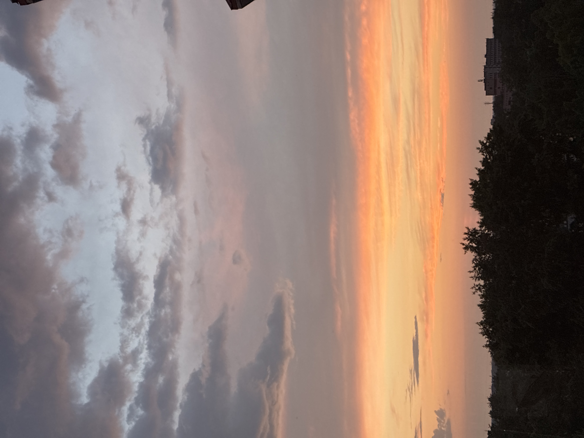 A wide-angle shot captures a twilight sky filled with soft hues of orange, pink, and purple, with streaks of grey clouds interspersed throughout. The horizon is lined with a dark silhouette of dense trees, and beyond them, a distant building is visible, bathed in the muted light. The scene appears to be a suburban or rural area, judging by the abundance of greenery and the absence of prominent urban structures. The overall mood is serene and tranquil, evocative of the end of the day.