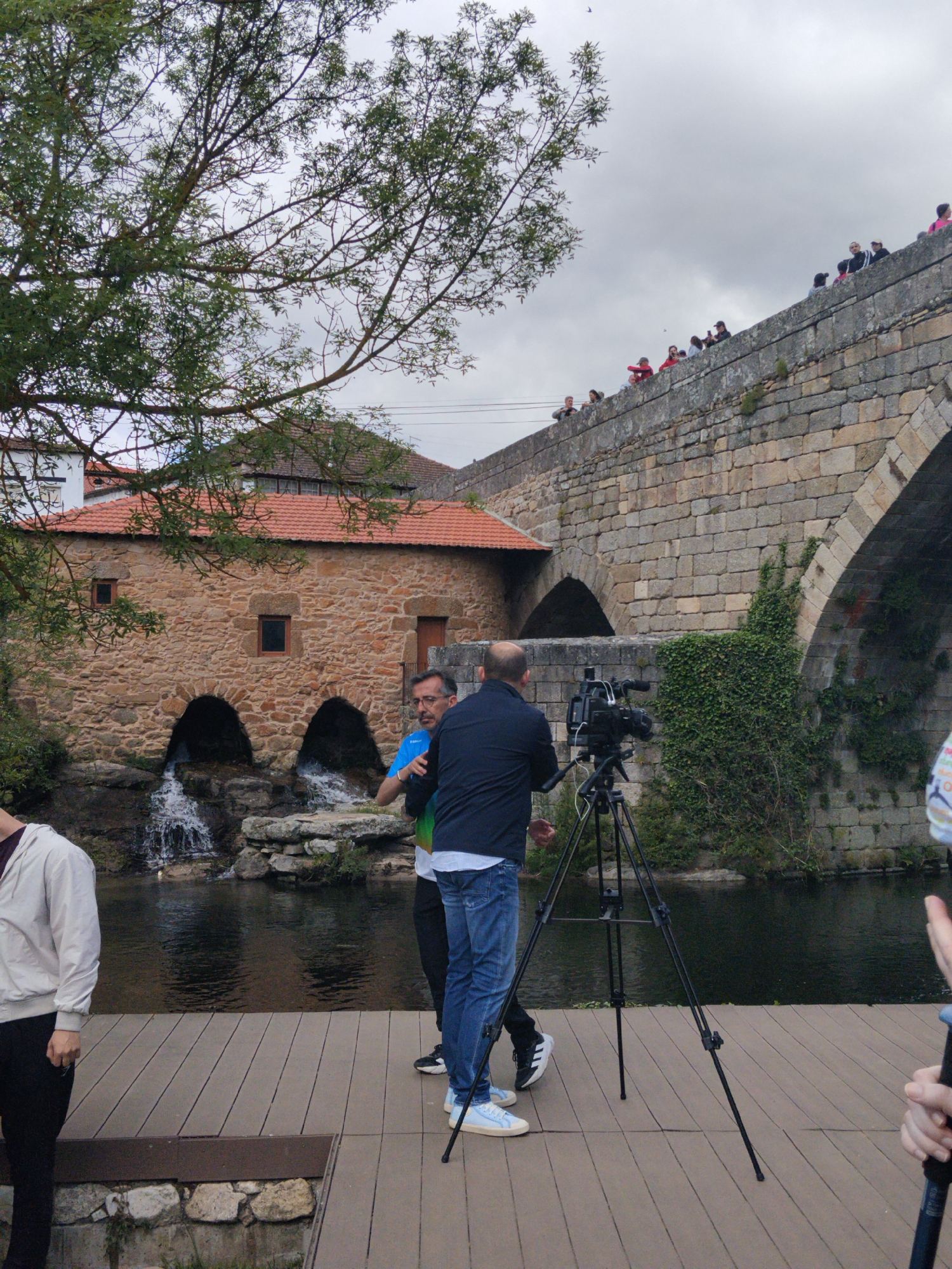 The image shows a scene in Tarouca, Portugal, featuring a stone building with a red tile roof, situated next to a river. The building has two arched openings at water level through which water flows, forming small waterfalls. A stone bridge with multiple arches spans the river; people are standing on top of the bridge, looking down. Two men are standing on a wooden platform on the riverbank, one facing the other. A professional video camera is set up on a tripod between them, suggesting they are involved in filming. Trees are on the left side of the image. The sky is overcast.
