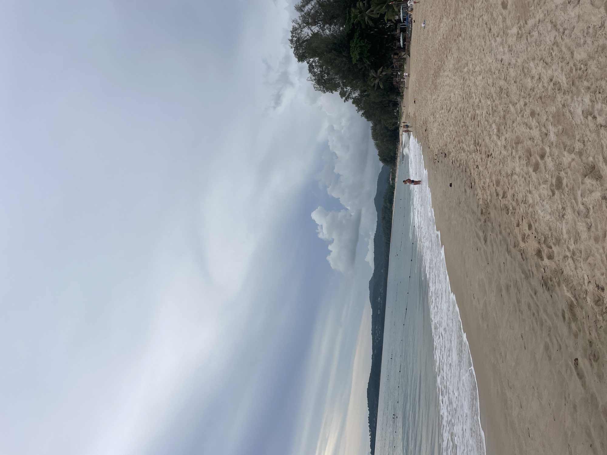 The image shows a beach scene in Choeng Thale, Thailand. The beach consists of beige sand meeting a light blue ocean, and a tree line on the right of the image. There are a few people on the beach and the clouds create a gray overcast sky.