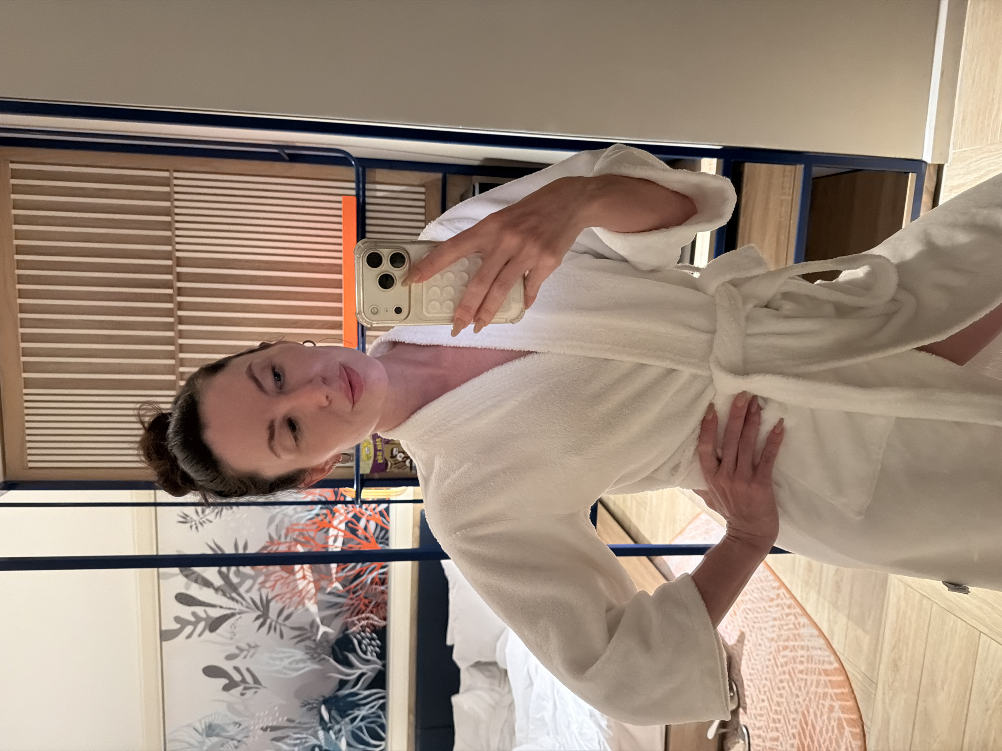 A woman is depicted in a mirror selfie taken within a room located in Dong Ninh Hoa Ward, Vietnam. She has dark hair styled in a high bun and is wearing a white terrycloth bathrobe, tied at the waist. Her left hand rests on her hip, while her right hand holds a light-colored smartphone with a transparent protective case, positioned to capture her reflection. The phone features a triple-lens camera system. Her nails are long and manicured with a light, natural-colored polish. The background, visible in the mirror, shows a room furnished with a bed featuring white bedding and pillows on the left. Behind the bed, a wall is decorated with a mural depicting marine flora in shades of grey, blue, and orange. To the right, a modular partition or shelving unit with dark blue metal frames and light wooden panels, some with vertical slat designs, is present. An orange horizontal stripe accents one of the wooden panels. The floor is covered with light wood-toned tiles, and a section of a light orange patterned rug is visible on the left side of the frame.