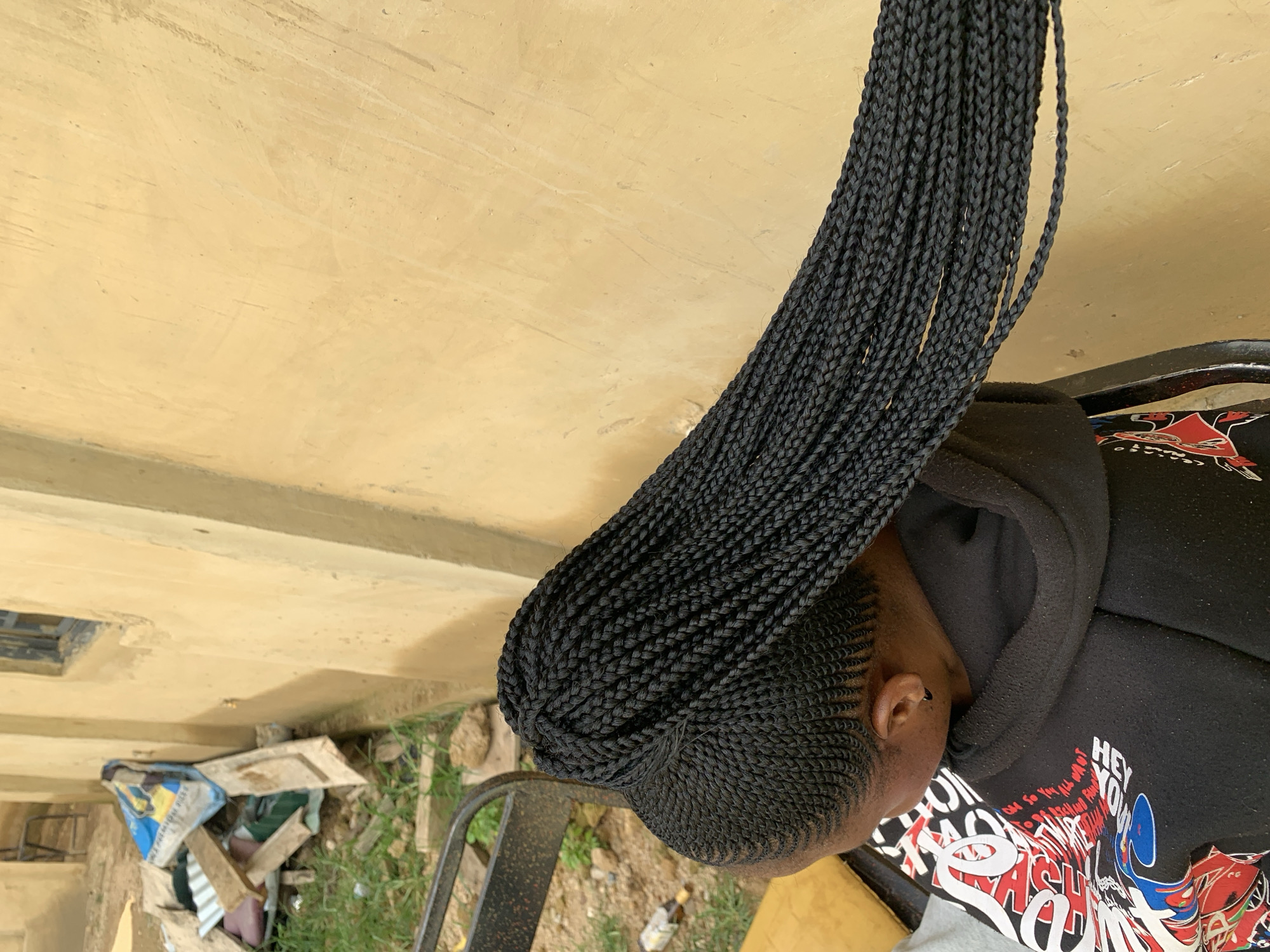 The image, taken in Jos, Nigeria, shows the back of a young woman's head and shoulders. She is wearing a black hooded sweatshirt with a colorful graphic print on the front. Her dark hair is styled in numerous thin braids that are gathered into a high ponytail, with the majority of the braids hanging down her back. The background is a textured, light yellow wall, possibly stucco. To the left of the wall, there is an area with scattered debris, including wood scraps and a blue and yellow bag, and some green grass and dirt. A dark metal chair is partially visible in the foreground.