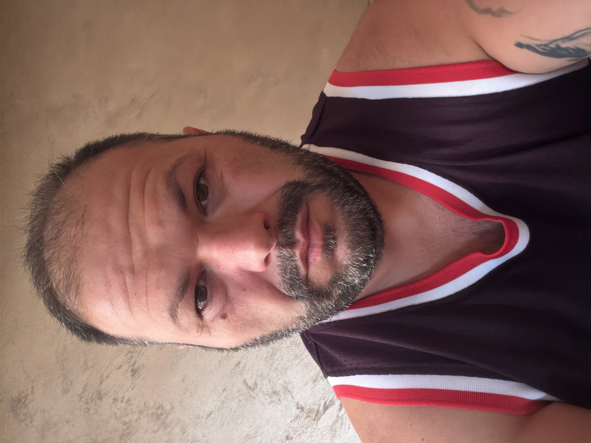 A medium shot portrait of a man with thinning dark hair, a full beard, and brown eyes. He is wearing a dark purple tank top with red and white trim. The background is a textured wall in a muted beige color. The image appears to be taken indoors, with natural lighting. There is no identifiable location in the city of Burgas, Bulgaria.
