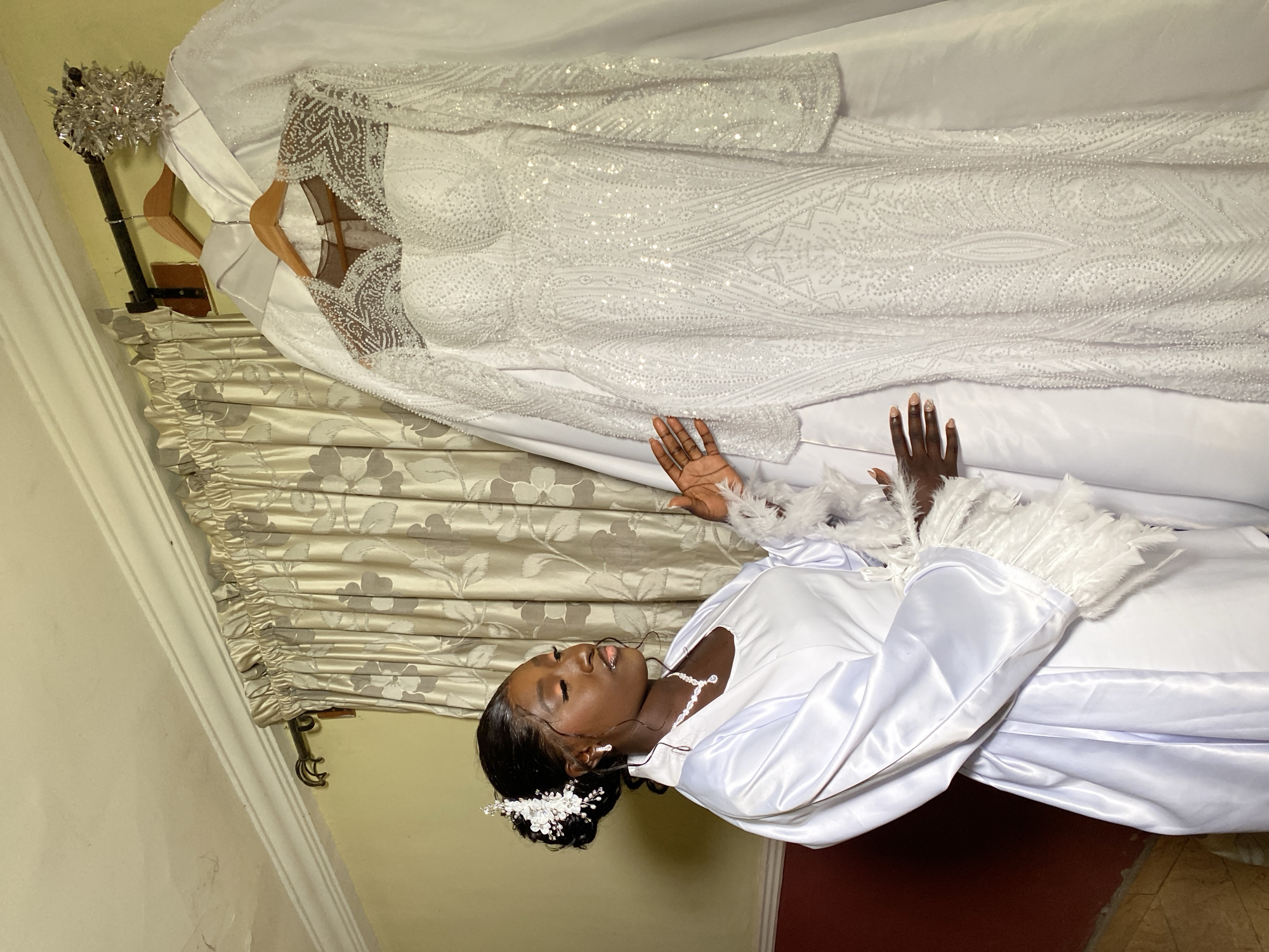 A young Black woman lies on her back, eyes closed, wearing a white satin robe with feathery trim, and a white floral hair accessory. She also wears a silver necklace and earrings. Above her hangs a wedding dress on a hanger, draped over a white sheet. The dress is white and heavily embellished with sequins. Behind the dress is a patterned, beige curtain. A wall is partially visible in the background. The location is presumed to be indoors, and the photograph may have been taken in Barikin Sabon Gidan Kanat, Nigeria. The woman is positioned as though she is admiring the wedding dress.