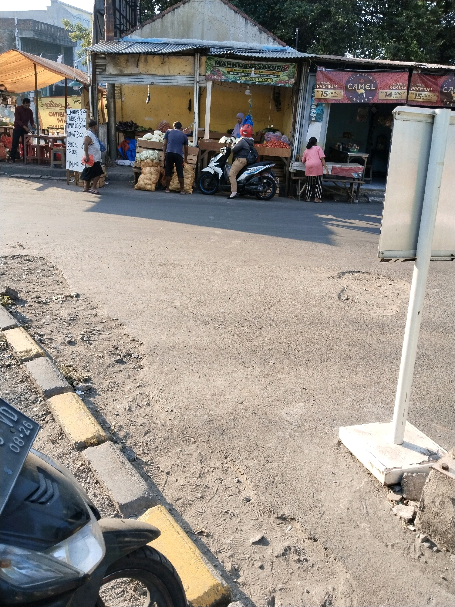 The image captures a street scene in front of small market stalls, likely in Wonocolo, Indonesia, on a sunny day. In the foreground, a black motorcycle is parked. The street is paved with asphalt and bordered by a curb with yellow and gray painted sections. To the left, market stalls are set up under a canvas awning. A man in a maroon shirt stands behind a table displaying goods. Further right, another stall features vegetables such as avocados and tomatoes. Two men, one in a blue shirt and the other in a dark shirt, are visible near piles of sacks. A woman in a white shirt and dark skirt walks by. A white motorcycle with a rider wearing a red helmet and brown pants is parked near the vegetable stall. Adjacent to this, a shopfront displays banners advertising "Camel Kretek" cigarettes with prices. A woman in a pink shirt and striped pants stands in front of this shop. A large, white, rectangular signpost stands on the right side of the image. The background shows buildings and trees, indicating an urban or semi-urban environment. Shadows suggest the sun is relatively high in the sky.