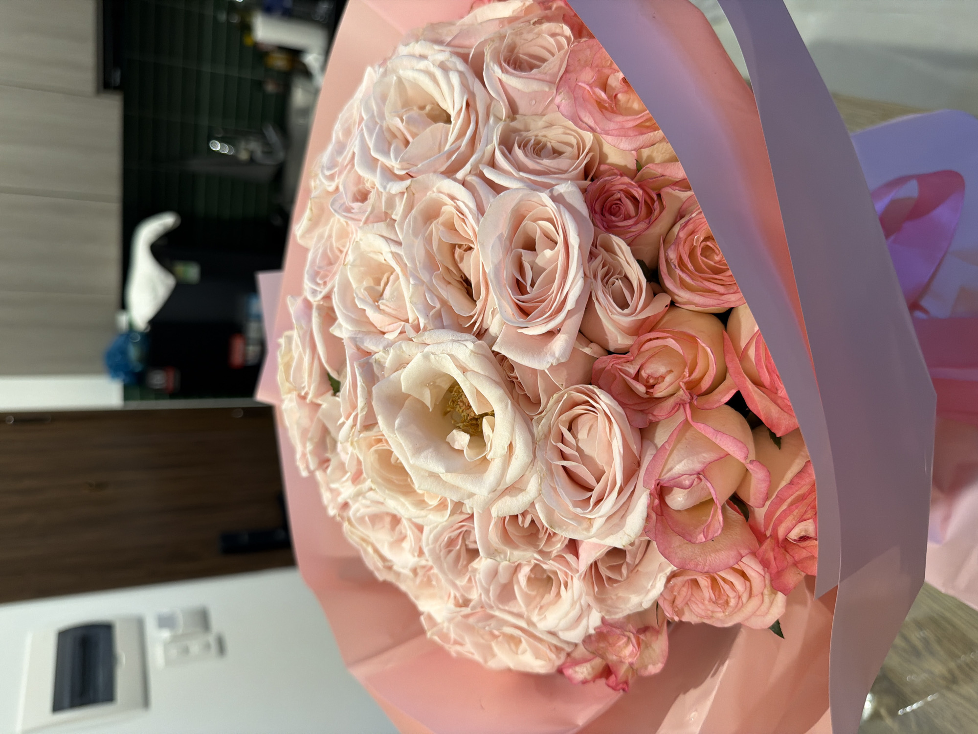 A close-up, eye-level shot focuses on a bouquet of pink roses, intricately arranged and partially wrapped in pale pink paper. The roses vary in bloom, with some tightly furled and others open, revealing delicate petals. The bouquet is positioned in the foreground, with a blurred background that suggests an indoor domestic setting. Visible elements in the background include a wooden door, a light-colored wall with electrical fixtures, and kitchen cabinetry with a tiled backsplash. The overall impression is one of a gift or decorative arrangement, likely in a residential area of Da Nang, Vietnam, given the context provided.