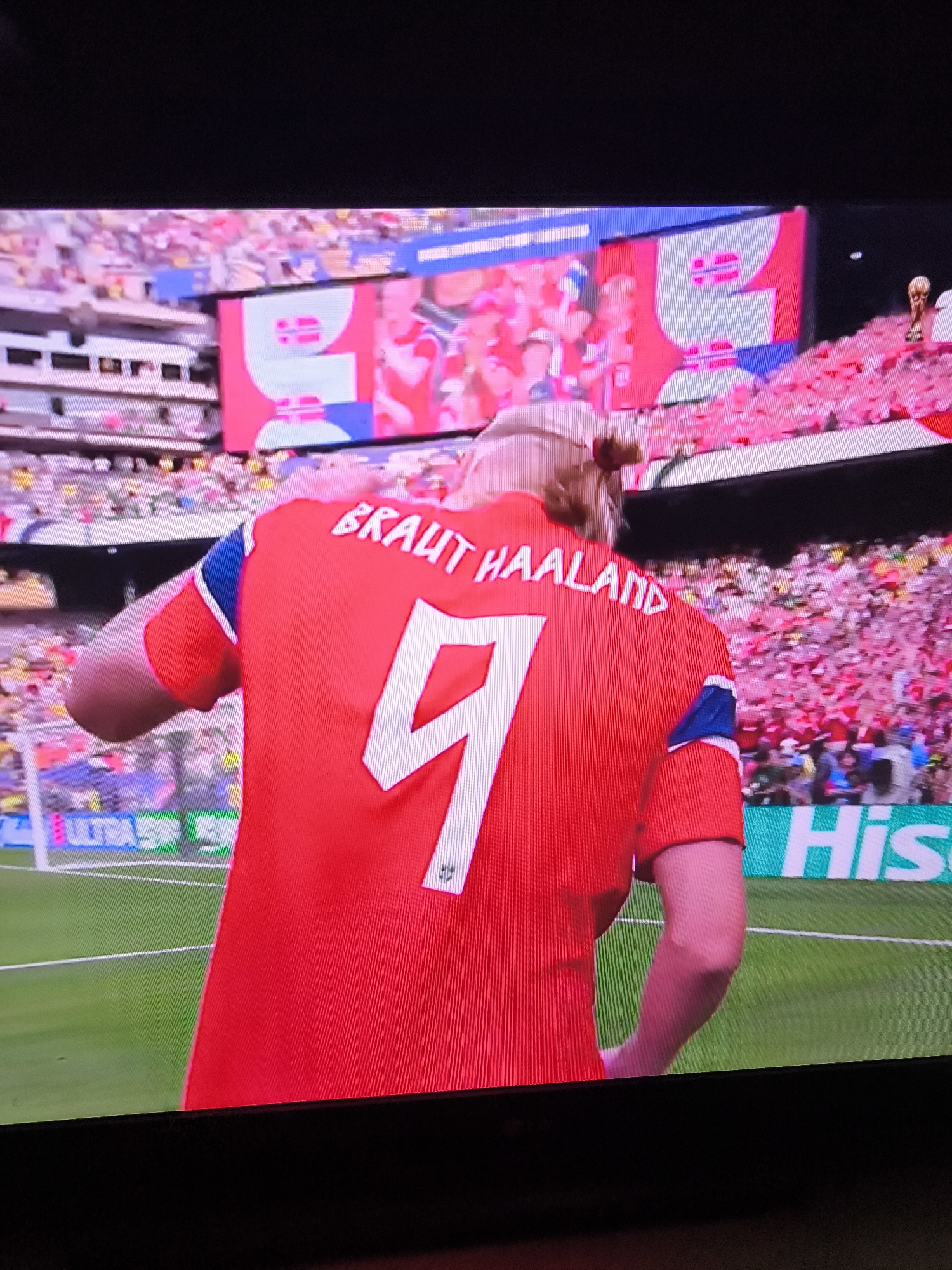 The image shows a rear view of a male athlete wearing a red soccer jersey with the name "BRAUT HAALAND" and the number "9" in white lettering. He has his hair tied up in a bun. He is standing on a green soccer field. Behind him, a large crowd of people in the stands are visible, many wearing pink. A large screen in the background displays graphics with red and white elements, and a trophy is visible on the right side of the screen. A green field with white markings is in the foreground. A portion of a sign with "His" in white letters on a green background is visible at the bottom right of the image. The setting appears to be a soccer stadium during a match or celebration.