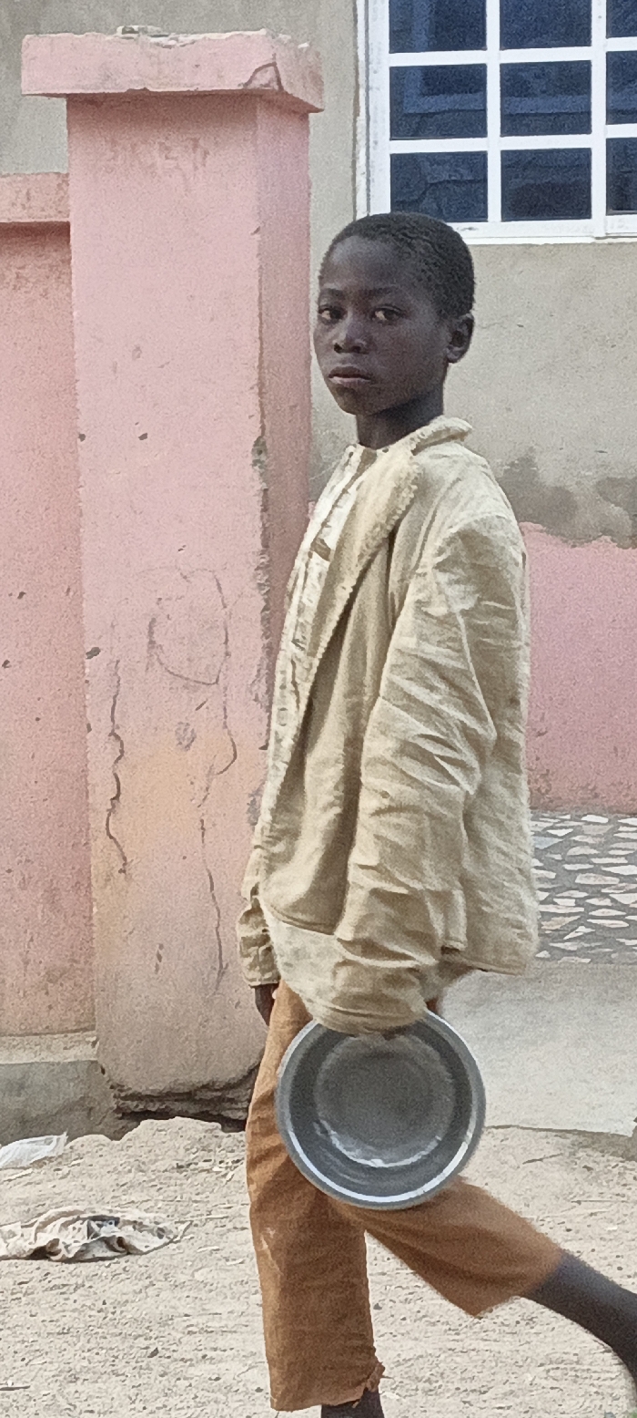 A young boy, likely of African descent, is the central subject, looking directly at the viewer with a serious or neutral expression. He has short, dark hair and is wearing a light-colored, possibly beige or tan, long-sleeved garment resembling a shirt or light jacket, which appears worn and wrinkled. He also wears brown trousers. In his right hand, hanging by his side, he holds a metallic, silver-colored bowl or pan.
The background features a textured, pinkish-orange wall showing signs of wear, including cracks and peeling paint. Behind it, to the upper right, is a window with prominent white frames and dark panes, set into a lighter, greyish wall. The ground is dusty and sandy, with some scattered debris on the lower left. Further back on the right, patterned paving stones are visible.
The scene takes place during the daytime under clear weather conditions. The boy's demeanor, his attire, and the presence of the bowl suggest he might be engaged in daily activities, possibly including begging or performing errands, in a setting characteristic of Rangaza, Nigeria. There is no visible text in the image.