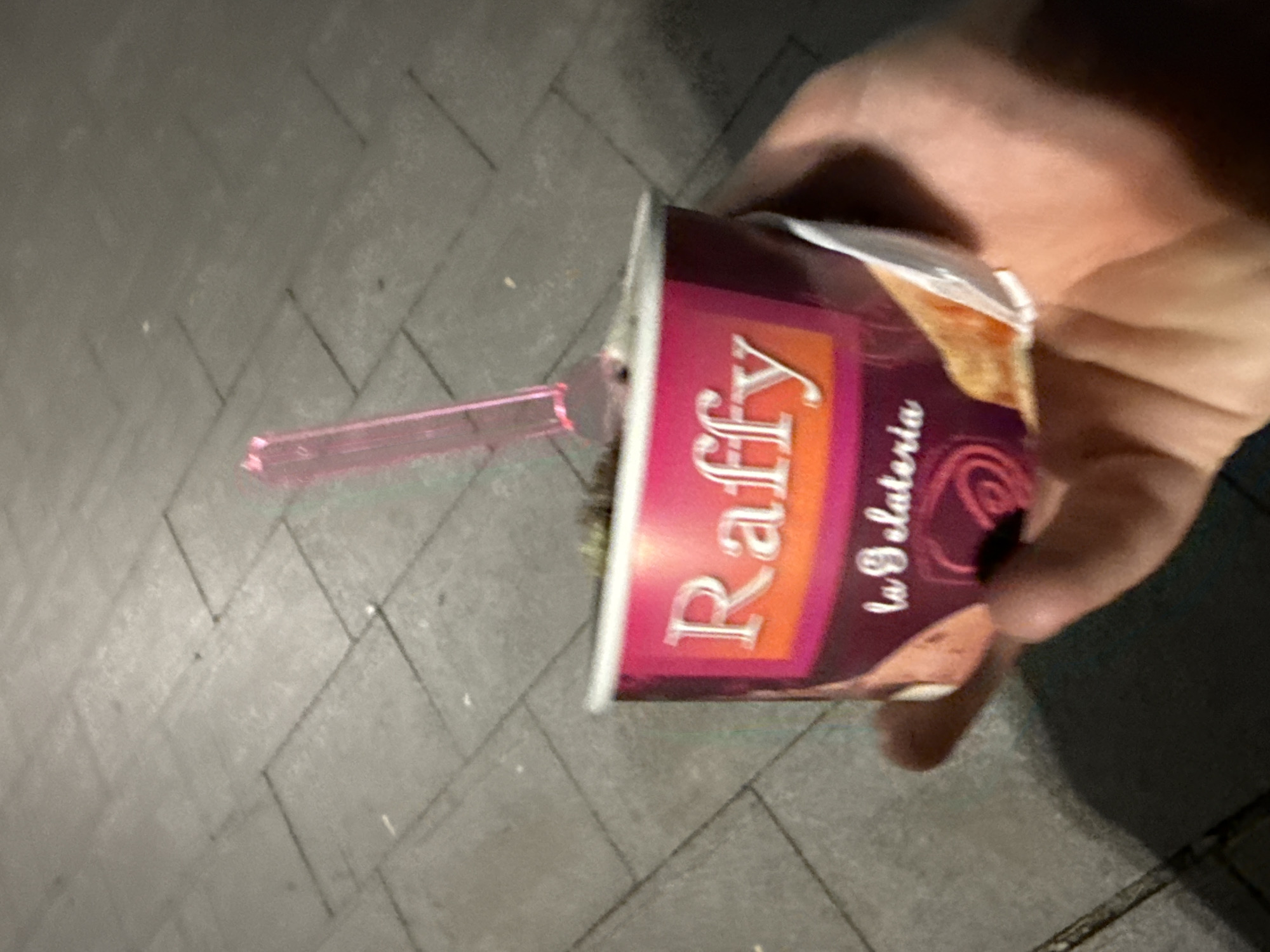 A hand is holding a paper cup of ice cream. The cup has the brand name "Raffy" and "la gelateria" printed on it. A pink plastic spoon is inserted into the ice cream. The background appears to be a sidewalk made of grey paving stones. The scene is likely in Stara Zagora, Bulgaria, given the context of the request.