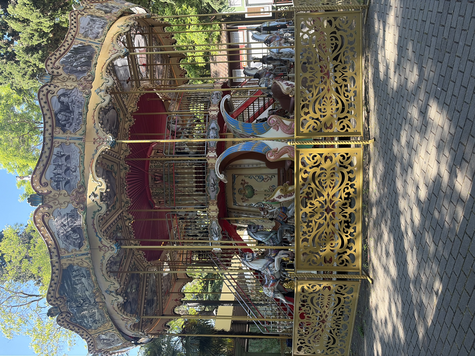 This media file showcases a carousel, a popular amusement ride, set in an outdoor location. The carousel is elaborately decorated with ornate gold metalwork, intricate carvings, and painted panels depicting various scenes, possibly historical or mythological. Several carousel horses, in different colors and designs, are visible, poised for riders. The carousel structure is multi-tiered, with red fabric canopies and a golden roof. A golden, decorative fence with intricate scrollwork is positioned in the foreground, partially obscuring the view of the carousel. The ground beneath is paved with gray interlocking stones, and dappled sunlight casts shadows, suggesting it is daytime, likely morning or afternoon, under clear weather conditions. Lush green trees and foliage surround the carousel, providing a natural backdrop. In the distance, a building with white and red elements can be seen. The scene appears peaceful and inviting, typical of a park or amusement area. No people are directly visible on the carousel or in the immediate vicinity, suggesting it might be closed or between rides. The location is identified as Sochi, Russia. There is no visible text in the image.