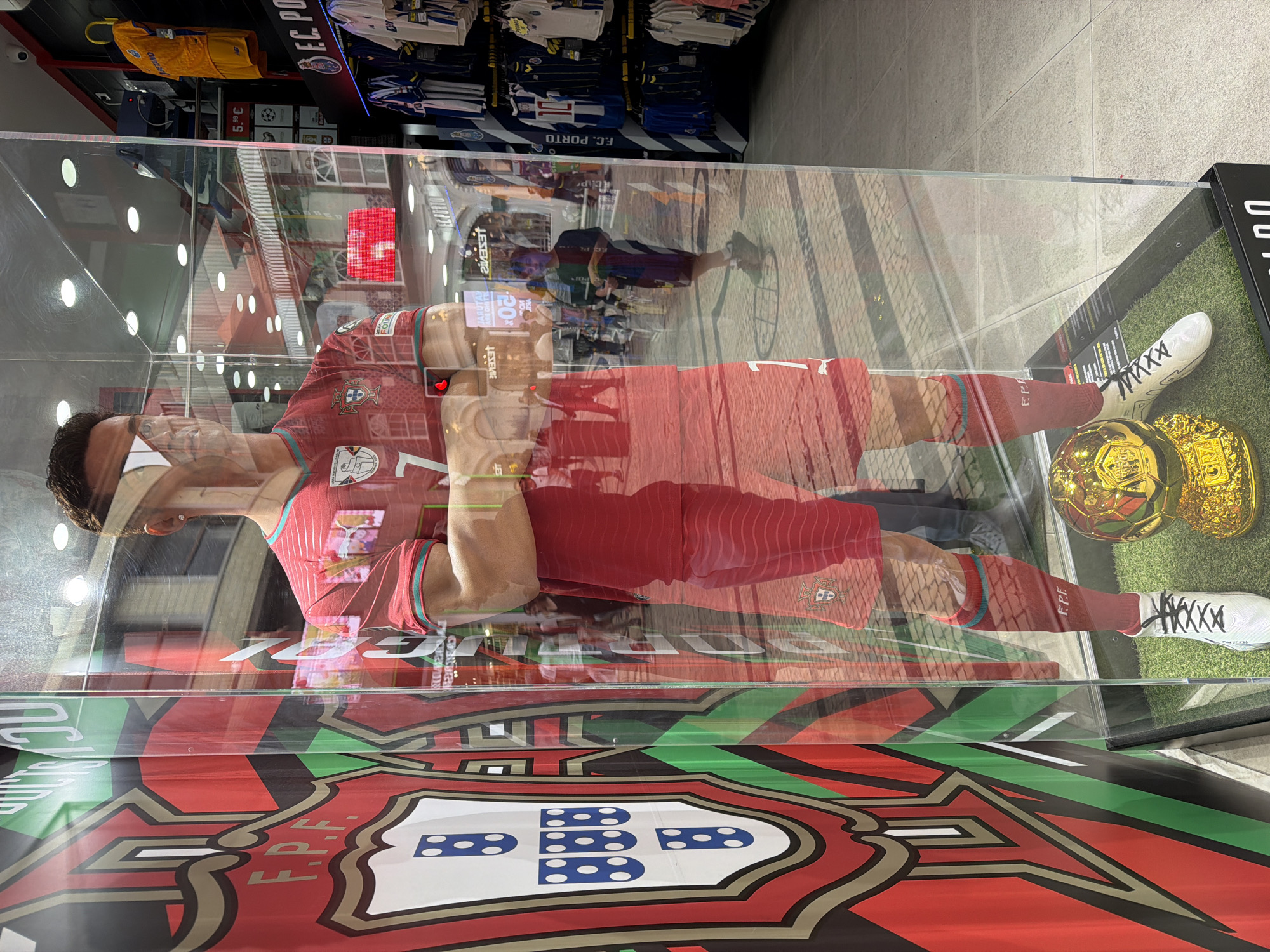 The image shows a display case located in Porto, Portugal, likely within a retail environment such as a sports store or souvenir shop. Inside the transparent case is a mannequin dressed in a red Portugal national football team kit with the number 7 on it. The mannequin is posed standing, with its arms crossed. Next to the mannequin's feet are two gold-colored football trophies and a pair of white football boots with black laces are also visible. The base of the display case features the Portuguese Football Federation (FPF) logo prominently displayed in red, green, white, and black. In the background, out-of-focus details show racks of football jerseys, including those of F.C. Porto. The store's interior is reflected in the glass of the display case, showing pedestrians walking on a paved surface. Signage and price tags are also visible in the background, along with lights and the reflection of the store's interior.