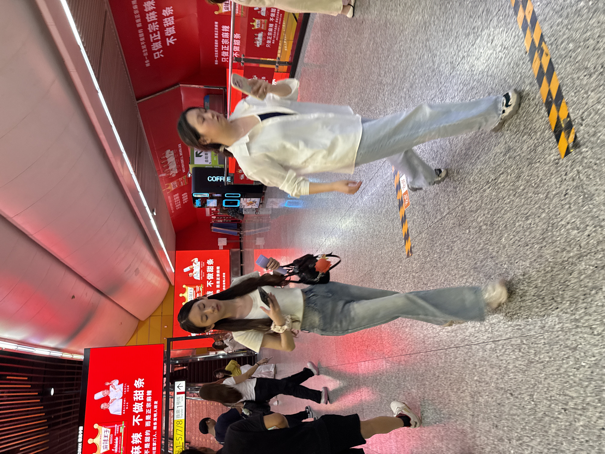 Here is a concise description of the scene:
The image shows an indoor scene, likely a public area such as a subway station in Yuzhong District, China. Numerous people are present, several walking. The scene is dominated by red and white advertisements with Chinese text. Some people are looking at their phones. A coffee vending machine is visible. The environment appears to be modern and urban. A yellow and black warning stripe is located on the floor.