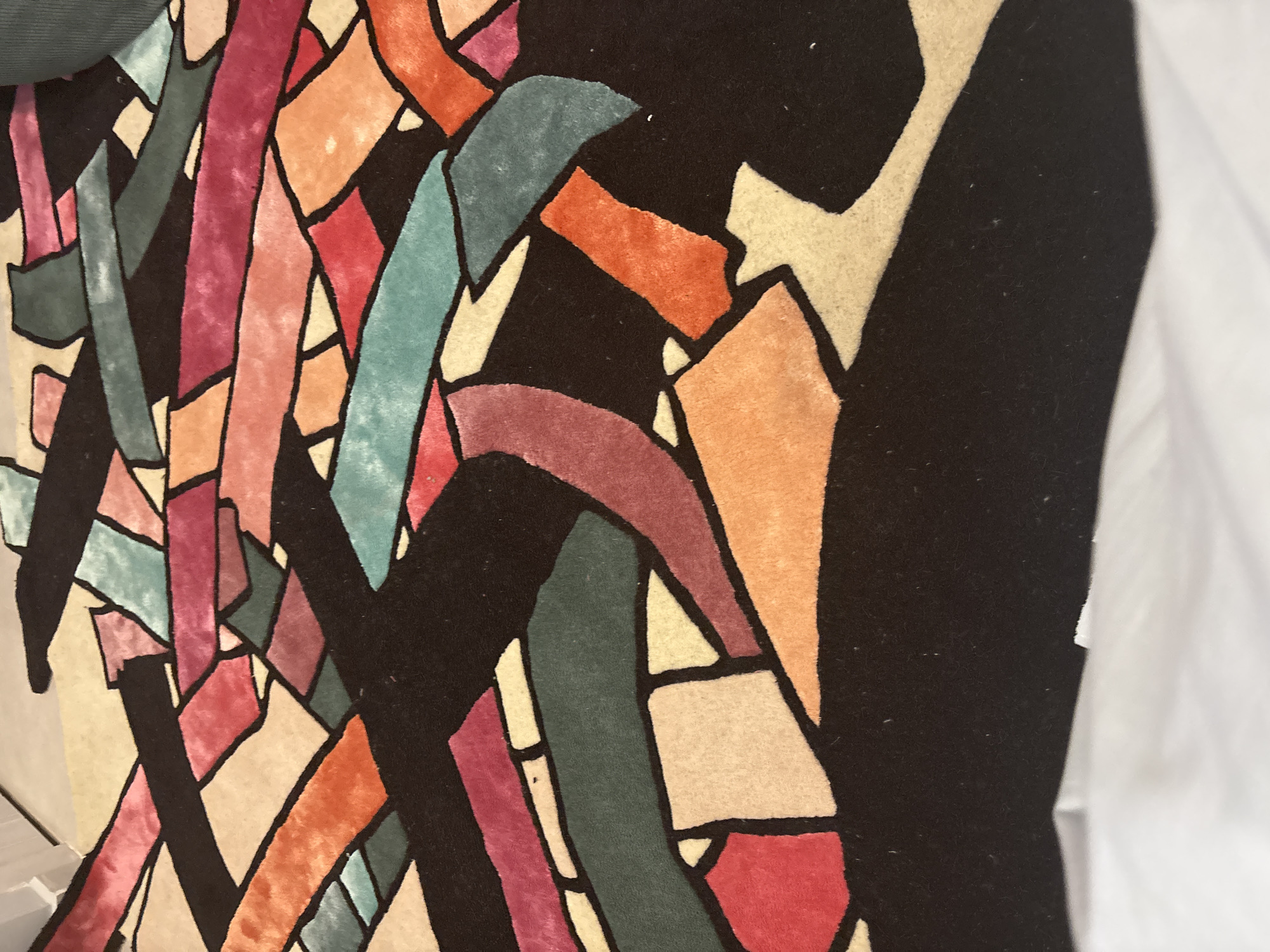 The image shows a close-up of a colorful textile with an abstract geometric design. The design features irregular shapes and lines in various colors, including shades of orange, pink, red, teal, green, and beige, outlined in black. The textile appears to be a rug or a decorative pillow. The setting is unknown but the mention of Arinj, Armenia is irrelevant to the content of the image itself.