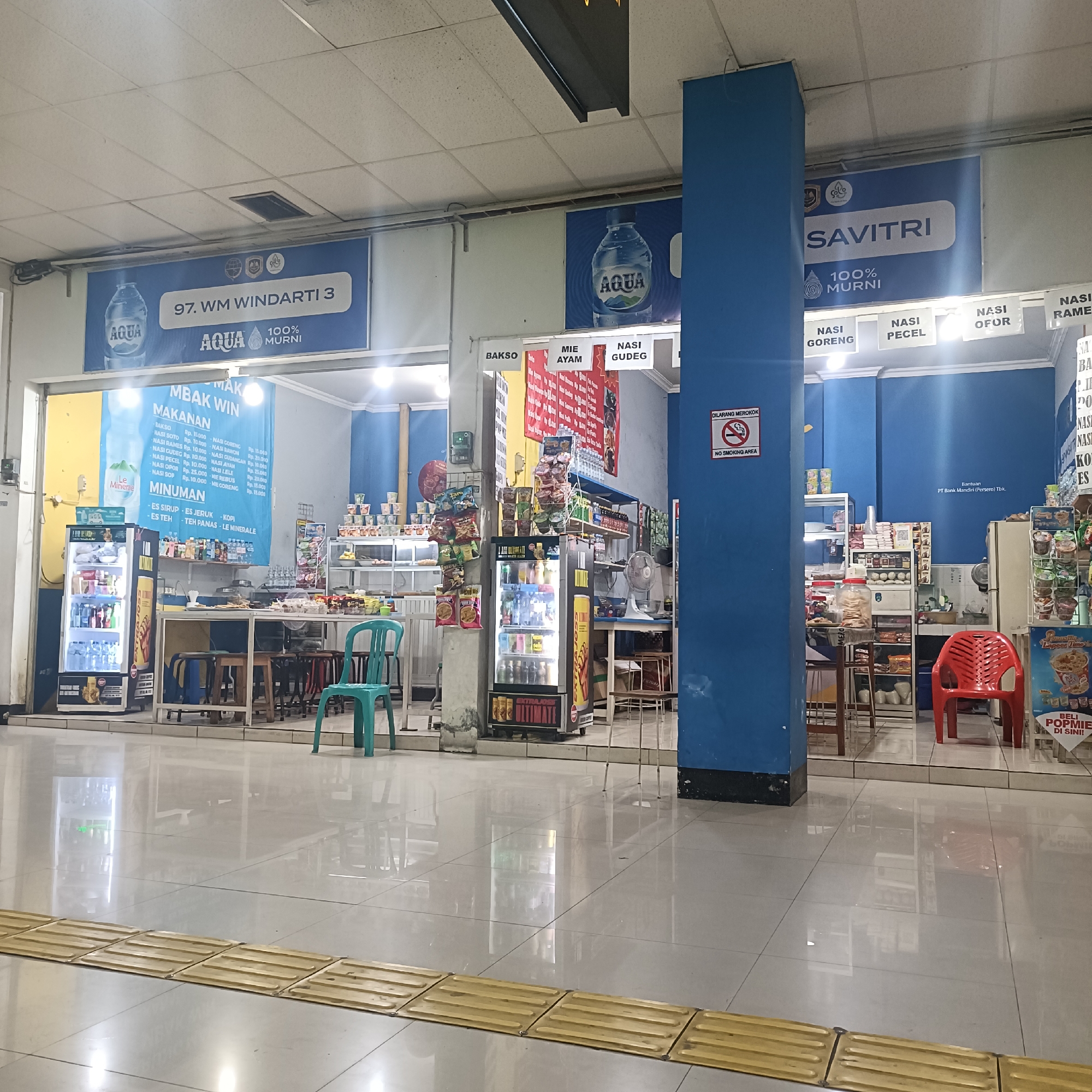 The image depicts an indoor marketplace or food court area, likely within a transportation hub, given the presence of signage and the clean, tiled flooring. The environment is well-lit, with a combination of overhead fluorescent lights and illuminated displays from refrigerators.
The foreground shows a yellow tactile paving strip, commonly found in public spaces to assist visually impaired individuals. Beyond this, the floor is a polished, light-colored tile, reflecting the surrounding environment.
In the mid-ground, there are several food stalls or small shops. On the left, a brightly lit refrigerator displays various beverages and food items. Next to it, a blue sign with white text announces "MBAK WIN MAKANAN MINUMAN," indicating it's a food and drink stall. This sign lists various Indonesian dishes and drinks with prices. A turquoise plastic chair is placed near this stall.
Further to the right, other stalls are visible, one with a sign for "BAKSO," "MIE AYAM," and "NASI GULUNG." Another stall features a cooler filled with drinks and shelves stocked with packaged food items, including instant noodles and snacks.
A prominent blue pillar is located in the center-right of the image, separating different sections of the marketplace. Above the stalls, large blue signs with white text are displayed. One sign reads "97. WM WINDARTI 3 AQUA 100% MURNI." Another sign, partially visible, has "SAVITRI" in large letters. Below the "SAVITRI" sign, smaller signs list food items like "NASI GORENG," "NASI PECEL," and "NASI OFOR." A "DILARANG MEROKOK" (No Smoking) sign is visible on the blue pillar.
In the background, more shelves are stocked with goods, and a red plastic chair is placed near a stall. The overall impression is a busy, functional commercial area.
While the image itself does not explicitly state the location, the mention of "Surakarta" as a potential location suggests that this marketplace is within that city in Indonesia. The types of food advertised (e.g., Nasi Goreng, Bakso, Mie Ayam) are common in Indonesian cuisine.