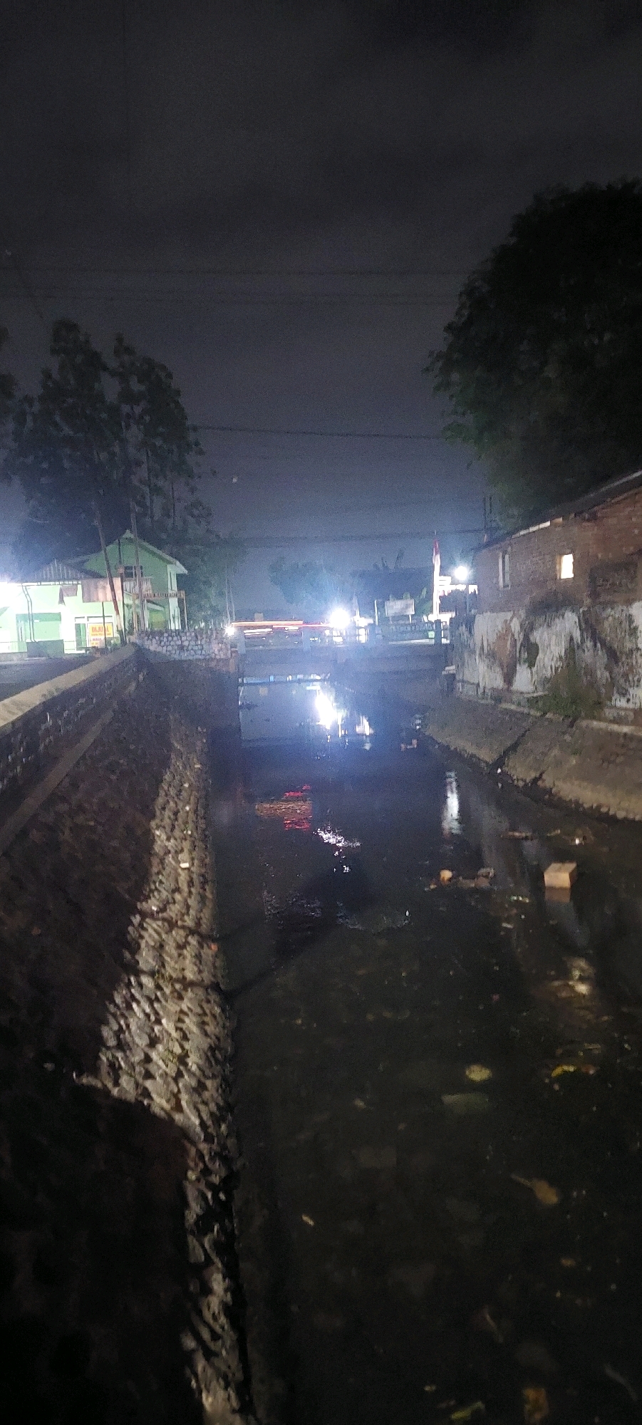 The image depicts a canal at night in Lumajang, Indonesia. The canal is lined with stone on the left bank and a brick building on the right bank. The water in the canal appears dirty and contains floating debris. Bright lights illuminate the scene from bridges and buildings, casting reflections on the water's surface. Trees and a green building are visible in the background, along with what appears to be a flagpole with an Indonesian flag. The sky is dark, indicating nighttime.