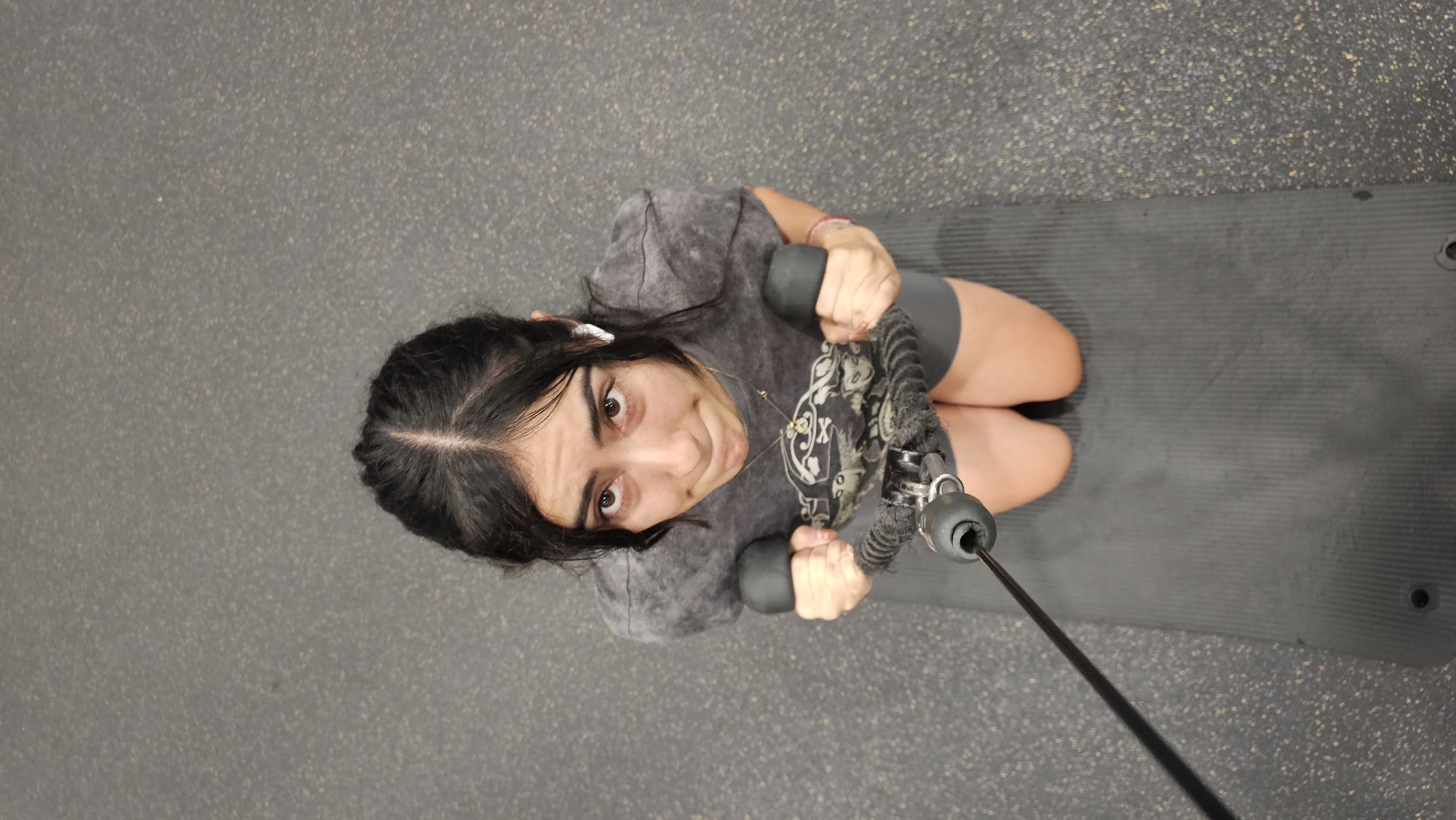 An extreme high-angle shot captures a young woman exercising in an indoor gym or fitness facility, located in Puebla, Mexico. She is positioned on a dark grey or black ridged exercise mat, which rests on a speckled dark grey gym floor. The woman has dark hair styled with a central parting, dark eyes, and a medium complexion. She is wearing a grey tie-dye t-shirt featuring a graphic design with stylized skull-like elements and unclear text, along with dark shorts or athletic wear. White earbuds are visible in her ears, and she wears a necklace with a small pendant.
She is actively engaged in a resistance training exercise, holding the black handles of what appears to be a cable row machine. A thick, braided black cable extends from the equipment she is holding towards the foreground. Her body position, with bent knees and engaged arms, suggests she is in the process of performing a seated row or a similar pulling movement. She looks directly up at the camera with a wide-eyed, playful expression, her mouth slightly contorted in a smirk or grimace, conveying a sense of surprise or effort during her workout. The lighting is consistent with an indoor setting.