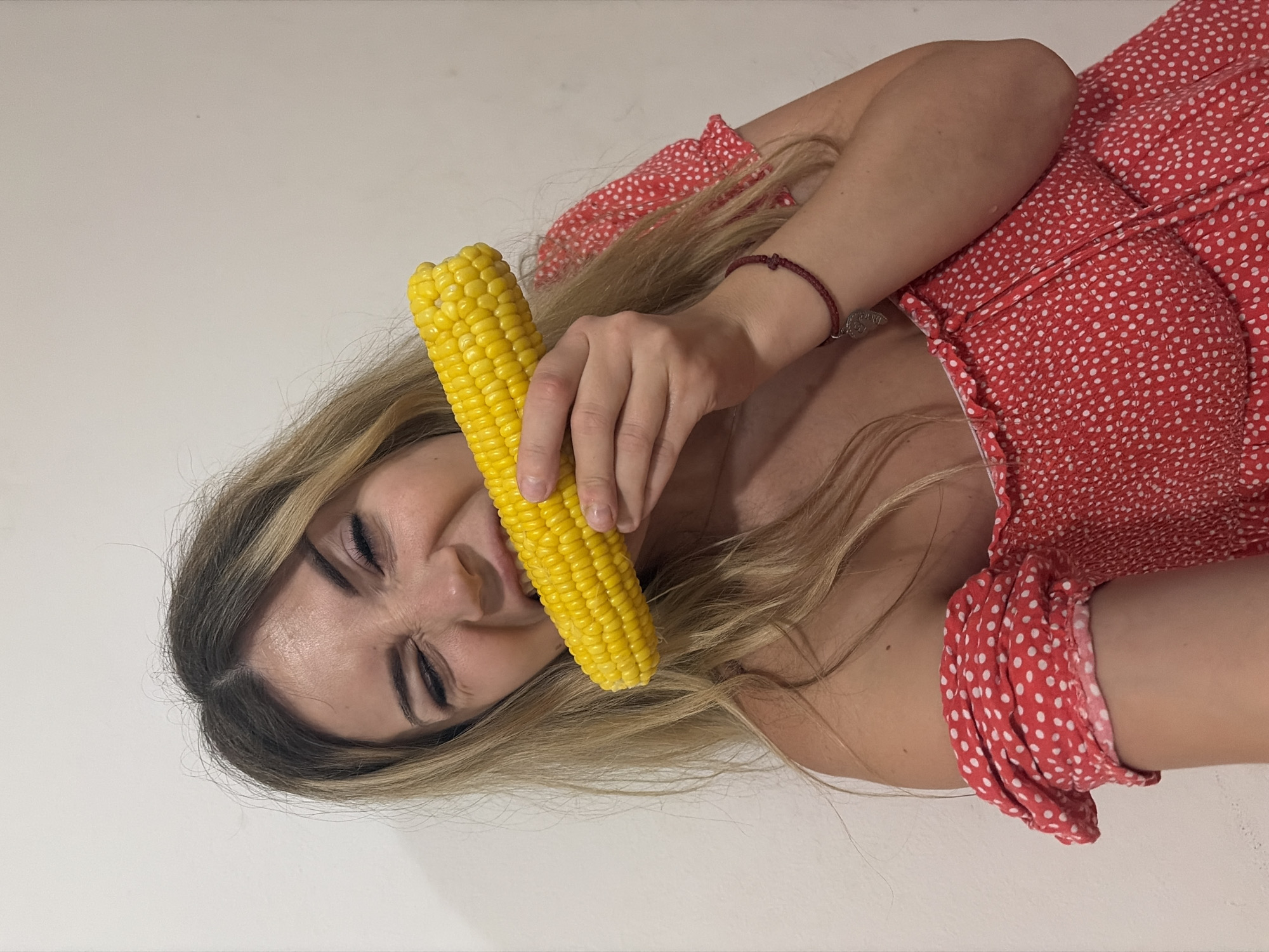 A blonde woman is biting into a cob of corn. She is wearing a red and white polka dot off-the-shoulder dress. A bracelet with a heart-shaped pendant is visible on her left wrist. She is standing in front of a plain, light-colored wall. The scene appears to be indoors.