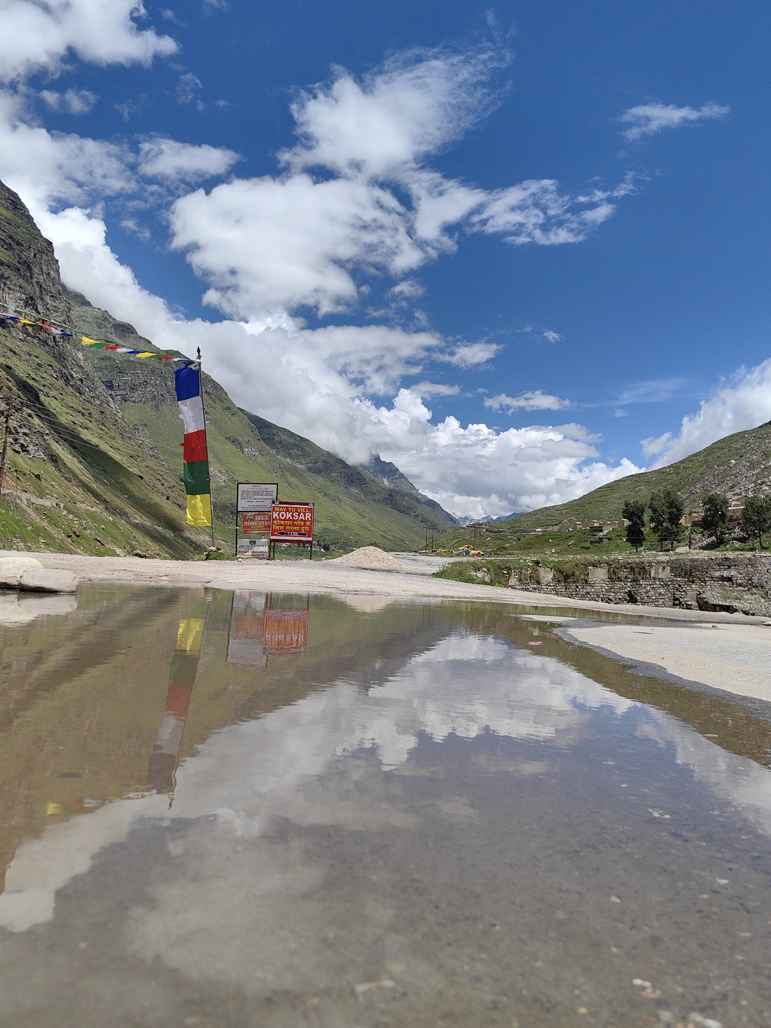 This is a low-angle, wide shot of a reflection of prayer flags and a sign in a puddle of water on a road in Koksar, India. The prayer flags are multicolored and hang on a pole. The sign is red and white and has text on it. The puddle reflects the flags, the sign, and the sky, which is blue with white clouds. The road is made of asphalt and gravel. In the background, there are mountains with green slopes. The scene appears to be a mountainous area in India.