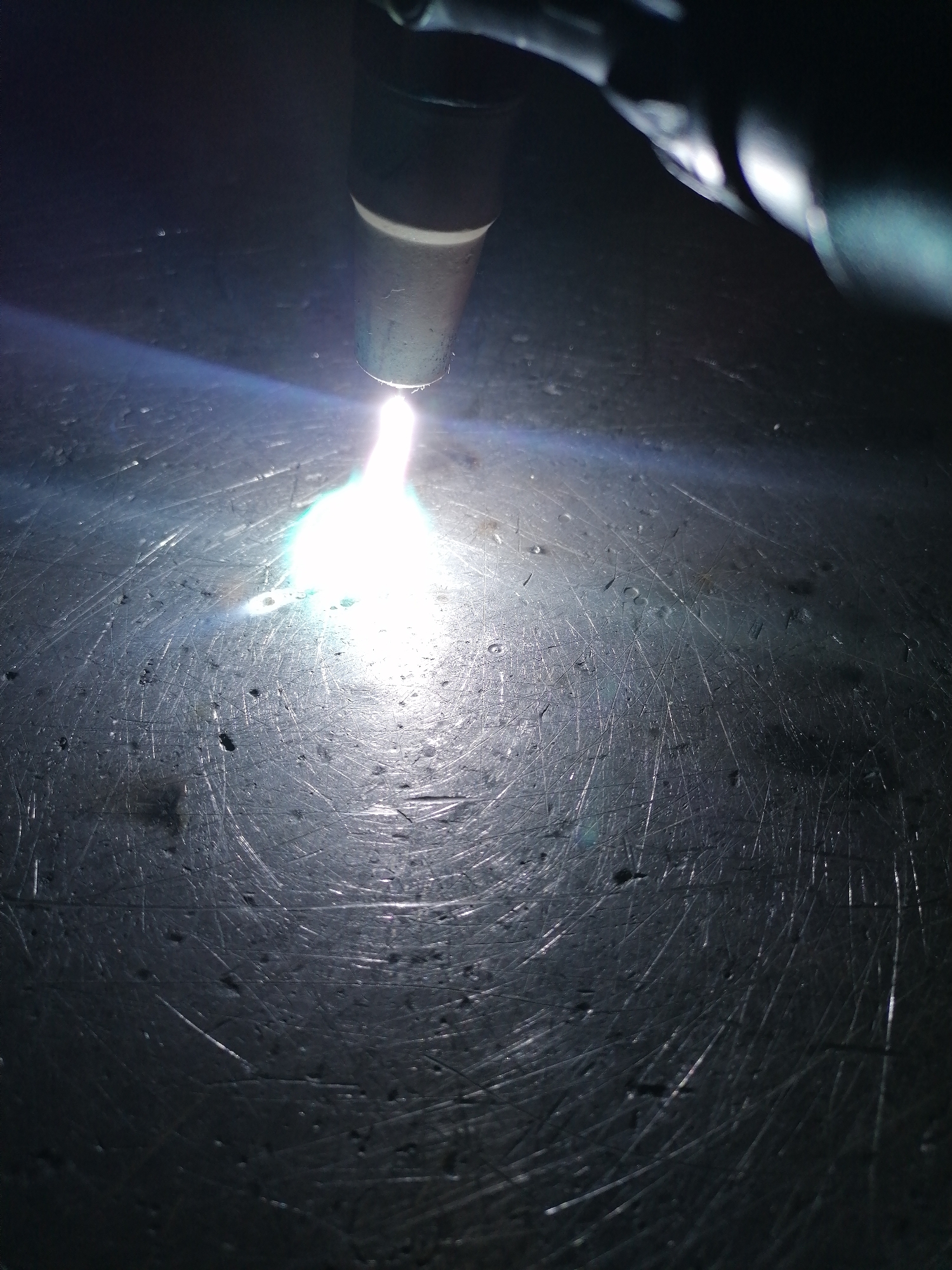 The image captures a close-up, high-contrast view of an industrial process, likely welding or plasma cutting, taking place in a dimly lit environment. The main subject is the tip of a tool, possibly a welding torch, positioned in the upper center of the frame. From its tip, an intensely bright, blue-white arc of light bursts downwards, illuminating the immediate work area.
This powerful light source reflects off a dark, metallic surface below it. The surface is heavily textured with numerous fine scratches, abrasions, and minor imperfections, suggesting a well-used workbench or a piece of metal being fabricated. The strong illumination from the arc highlights these intricate details on the metal, revealing its worn character. The rest of the scene, particularly the upper background, is dark and out of focus, emphasizing the concentrated light and activity at the point of operation. There are no people visible, nor any discernible text. The overall impression is one of intense industrial work, focusing on the creation of high energy to shape or join materials.
