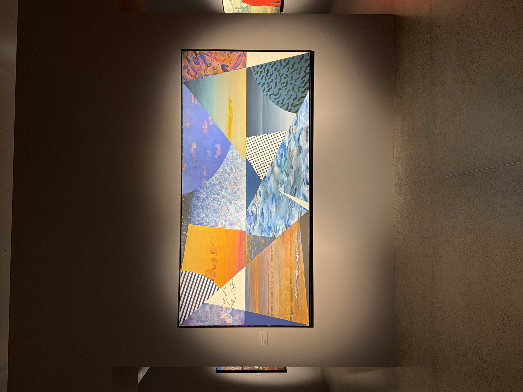 A tall, vertical, abstract painting hangs on a subtly textured, dark grey wall, illuminated by a warm spotlight from above. The painting is composed of numerous geometric shapes and patterns in a wide array of colors, including blues, purples, yellows, oranges, and white, with some areas featuring stripes, dots, or swirling brushstrokes. At the bottom of the painting, a small, white label is affixed to the wall. Below this, a second, much smaller, horizontally oriented framed artwork is visible, also illuminated by the spotlight, though its contents are indistinct. The overall setting appears to be an art gallery or exhibition space, with the lighting creating a dramatic and focused atmosphere. The lighting suggests an indoor environment, likely during evening hours or in a controlled lighting setup.