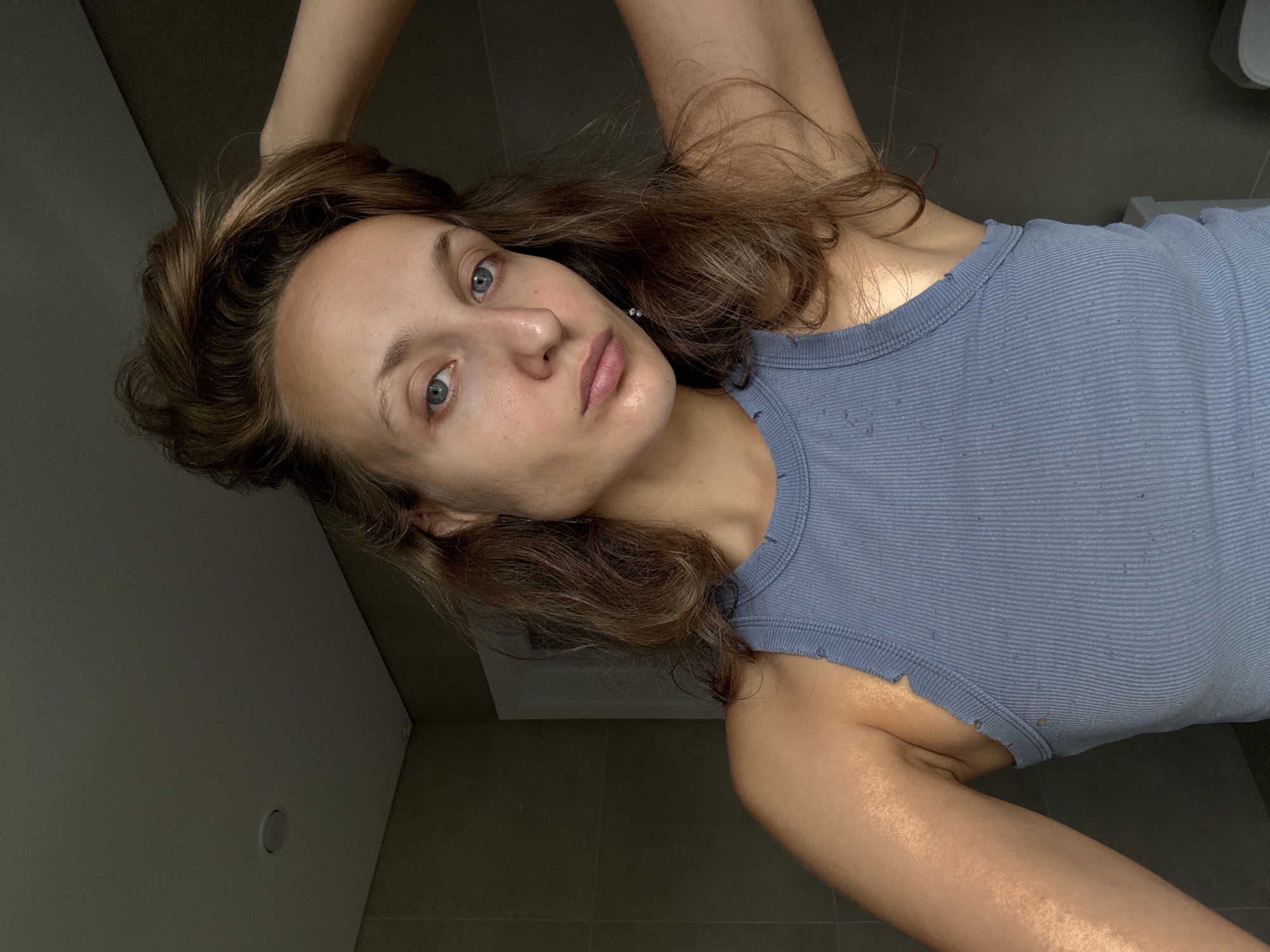 The image shows a close-up, selfie-style shot of a young Caucasian woman in a blue, ribbed tank top. She has brown hair styled loosely, with some strands falling across her face. Her head is tilted slightly, and her right hand is raised to the back of her head, pulling some of her hair up. She is looking directly at the camera with blue eyes. Her facial features include prominent cheekbones, a small nose, and full lips. Her skin appears bare, with some natural texture visible. The background is a dimly lit bathroom with gray tiled walls and a ceiling with recessed lighting. A door or cabinet is visible on the right side, along with what appears to be the top of a toilet tank. The lighting suggests an indoor setting, possibly natural light coming from a side window, creating highlights on her arm and shoulder. The scene is forensic in its detail, with no specific elements indicating a location in the city of Подшивалово, Russia.
