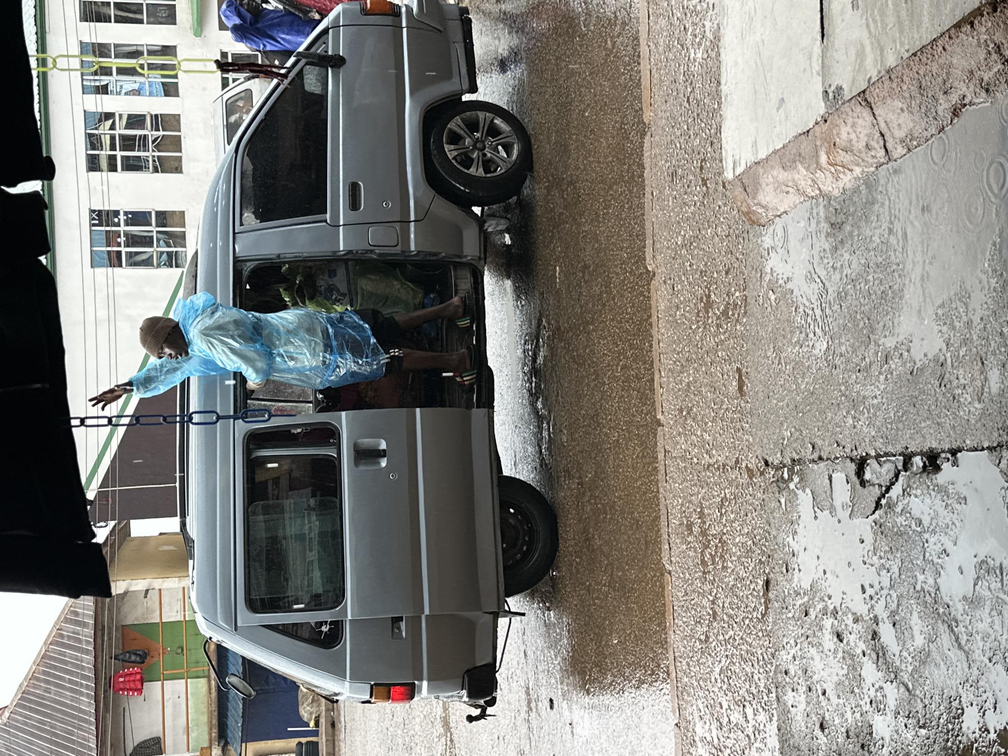 In Bukuru, Nigeria, during a downpour, a person stands partially inside a gray van, reaching out of the open sliding door. The individual, clad in a translucent blue plastic poncho and a tan beanie, gestures upwards with their right hand. Their legs are visible, wearing dark shorts with white stripes and multicolored slide sandals. The van's doors are open, revealing the interior and another person in the back. The van is parked on a wet, paved surface, with significant puddles reflecting the overcast sky and surrounding buildings. In the background, a multi-story building with numerous windows stands, and to the left, a structure with a corrugated metal roof displays hanging red and black items. The street is slick and filled with rainwater, indicating ongoing precipitation.