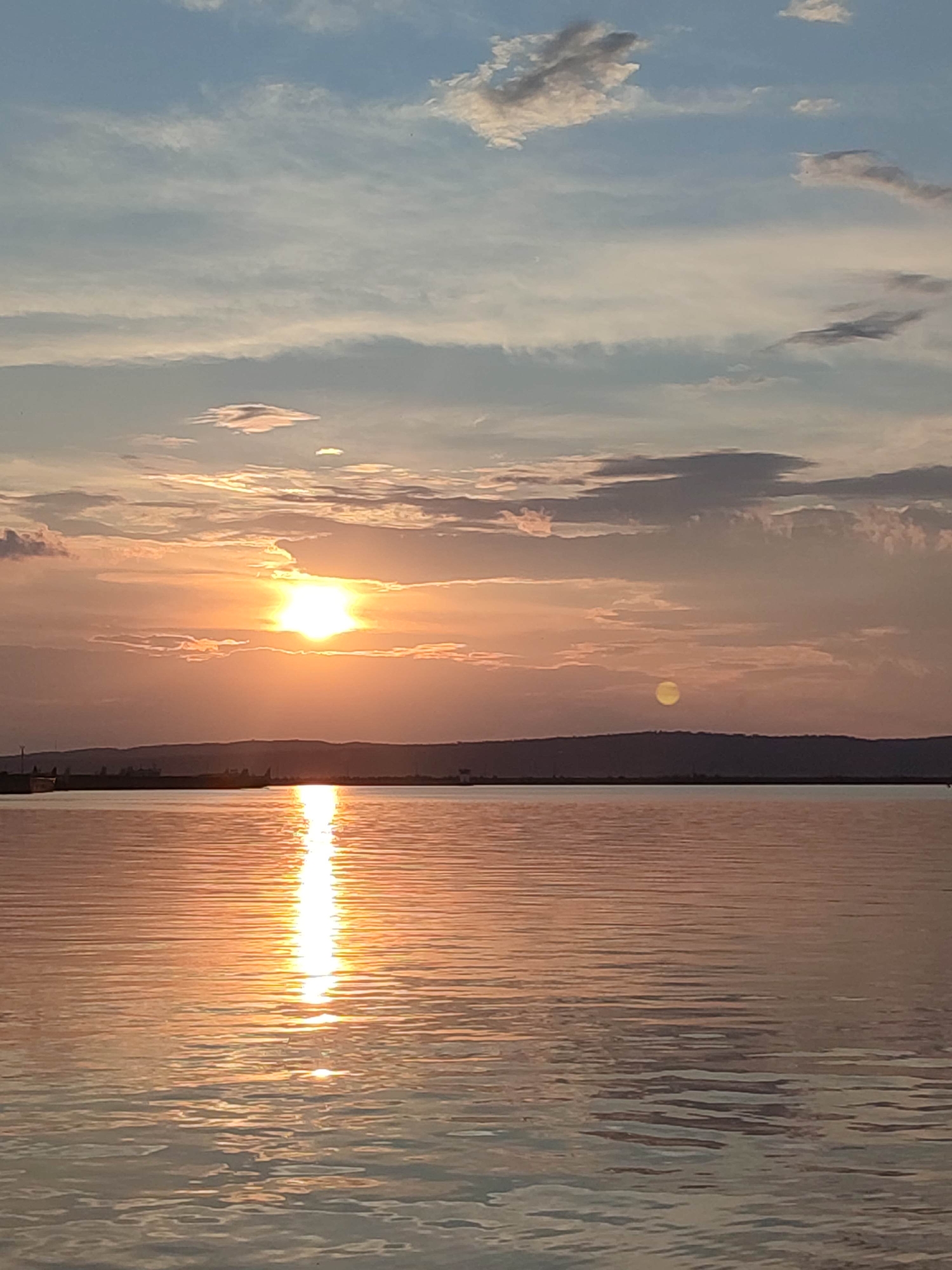 A sunset over a body of water is depicted. The sun, a bright orb, is low in the sky, casting a golden glow across the scene. Clouds are scattered across the sky, some tinged with orange and pink from the setting sun. A dark, hilly landscape forms the horizon line, with what appears to be structures or trees dotting its edge. The water in the foreground is calm, with gentle ripples reflecting the light from the sky. The overall mood is serene and peaceful. The image appears to be taken from a boat or the shore.