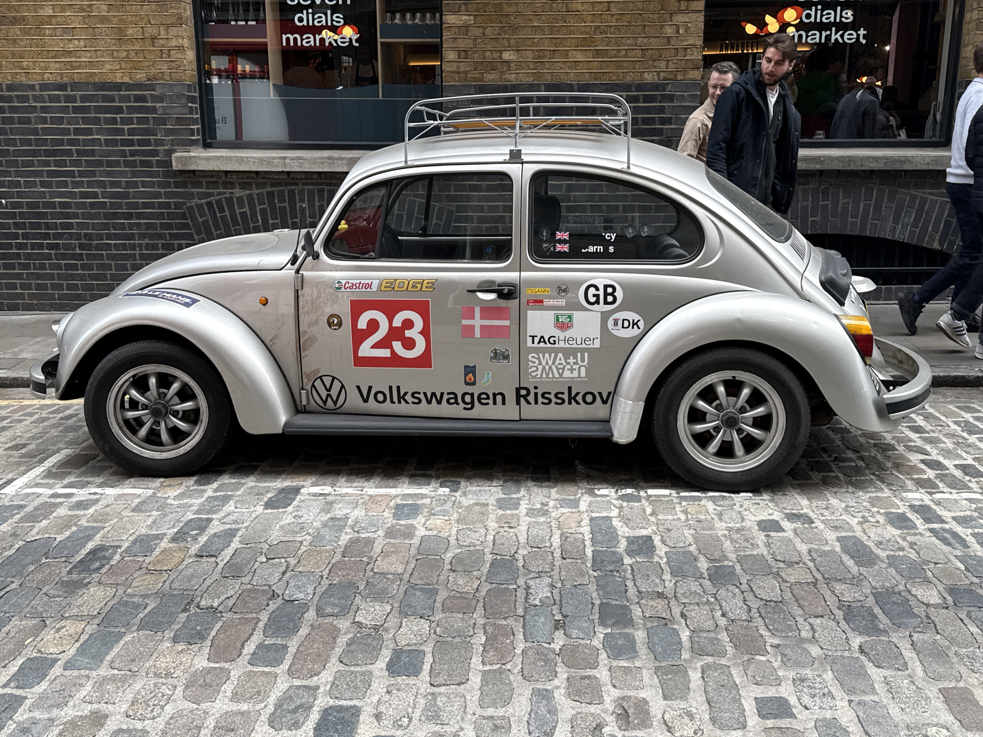 The image shows a silver Volkswagen Beetle parked on a cobblestone street. The car has a roof rack and several stickers, including a red "23" and flags of the UK, Denmark, and Germany. "Volkswagen Risskov" is written on the side of the car. In the background, there are people walking by and a building with the words "seven dials market" visible on a window. The image is taken in the Seven Dials Market area of London, United Kingdom.