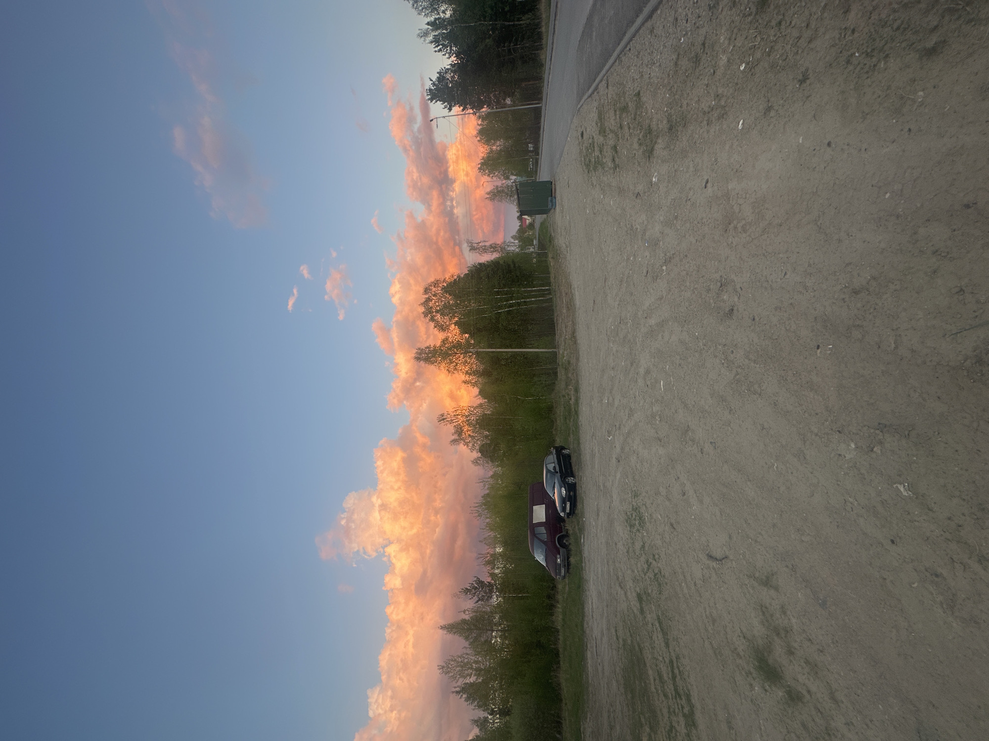 The image, correctly oriented, depicts a serene outdoor scene during either sunset or sunrise, characterized by a strikingly colorful sky. The upper left portion of the sky is clear blue, gradually transitioning to a vibrant display of orange, pink, and yellow clouds that stretch across the horizon on the right, bathed in the warm, soft light of the golden hour.
In the foreground, a wide, unpaved, dusty ground occupies the left side of the frame, interspersed with sparse patches of green vegetation. Two vehicles are parked on this ground: a dark-colored car and a maroon van, possibly a Mercedes-Benz Sprinter.
A dense line of trees, including both coniferous and deciduous types like birch, forms a prominent horizontal band across the middle ground, some appearing silhouetted against the bright sky. A paved road runs alongside the unpaved area, bordering the treeline. Near the road and trees, a dark green utility box or small shed is visible, with a streetlight pole extending upwards above the treeline.
No people are visible, and no specific activity is taking place beyond the presence of the parked vehicles. The scene conveys a tranquil, semi-rural or rural setting, consistent with the provided location context of Высокий, Russia. There is no visible text in the image.