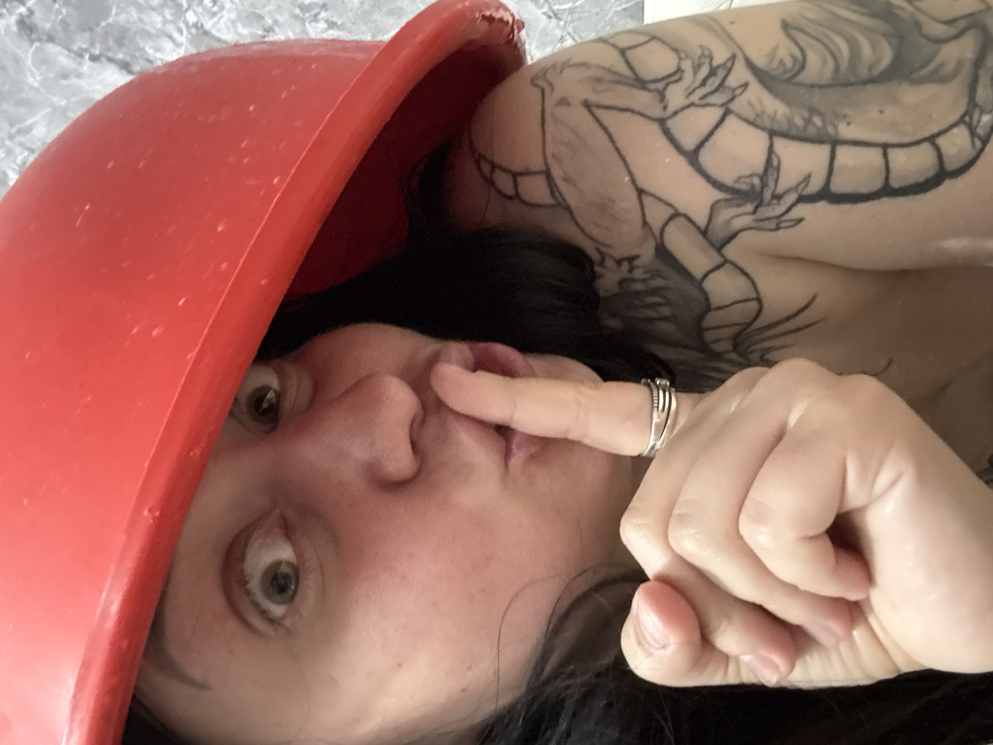 A close-up, rotated portrait features a woman looking directly at the camera from behind a large, bright red, bowl-shaped object. The red object, possibly a plastic basin or tub, occupies the left side of the frame, partially obscuring her face and dark hair. Small water droplets are visible on its surface. The woman has wide, light-colored eyes, conveying a playful or surprised expression. Her right index finger is pressed against her nose or upper lip, and a silver-toned, multi-banded ring is worn on another finger of her right hand. A prominent black and grey tattoo, depicting a creature with claws and a segmented body (possibly a dragon), is visible on her right shoulder and upper arm. The background is a mottled light gray and white, resembling a tiled wall, suggesting an indoor setting. There is no visible text.