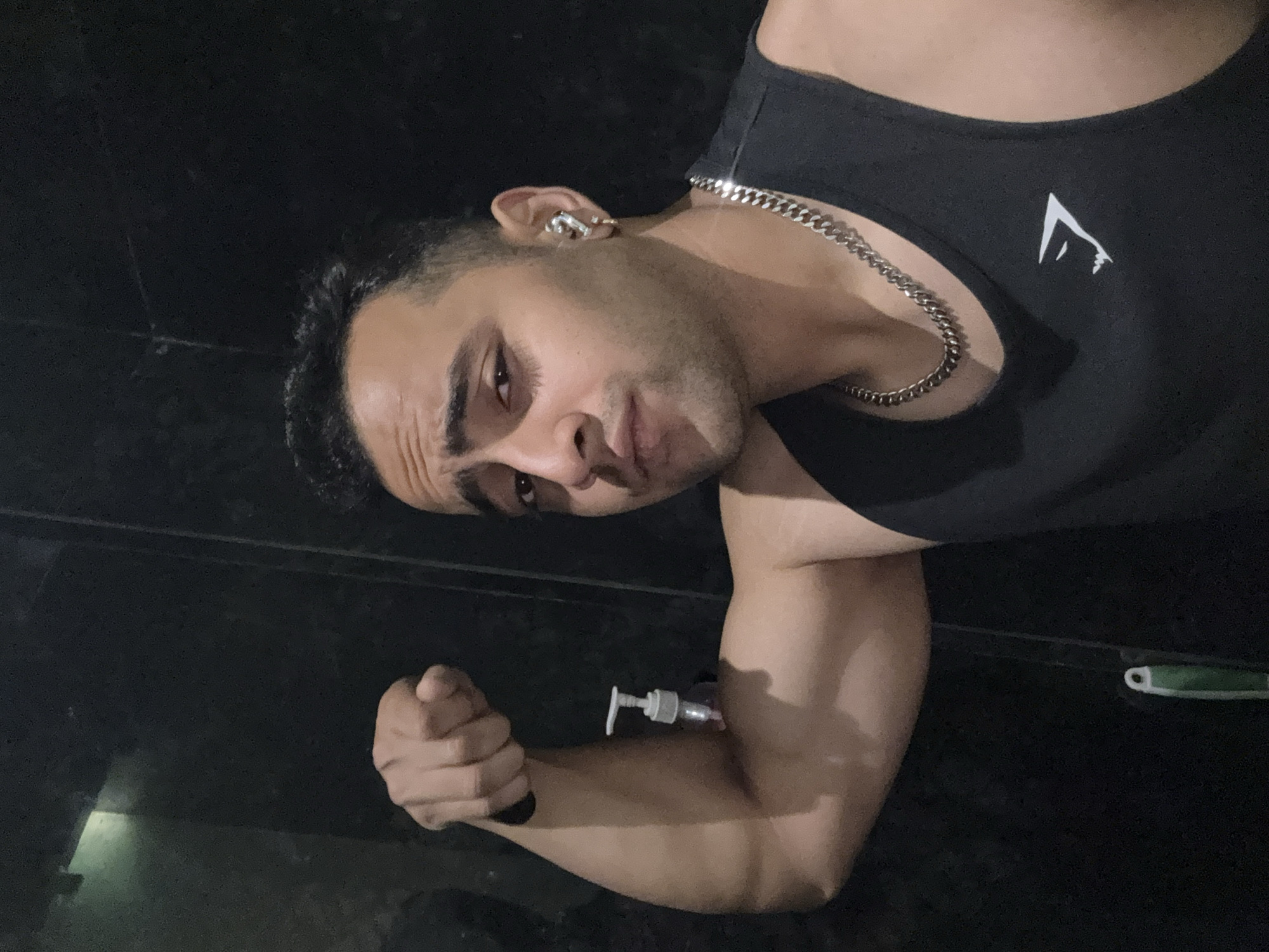 A person in a black tank top with a white logo and a silver chain is flexing their bicep, with a soap dispenser held between their arm and body. They also have a silver earring. The location is not discernible as Kolkata, India based on the image.