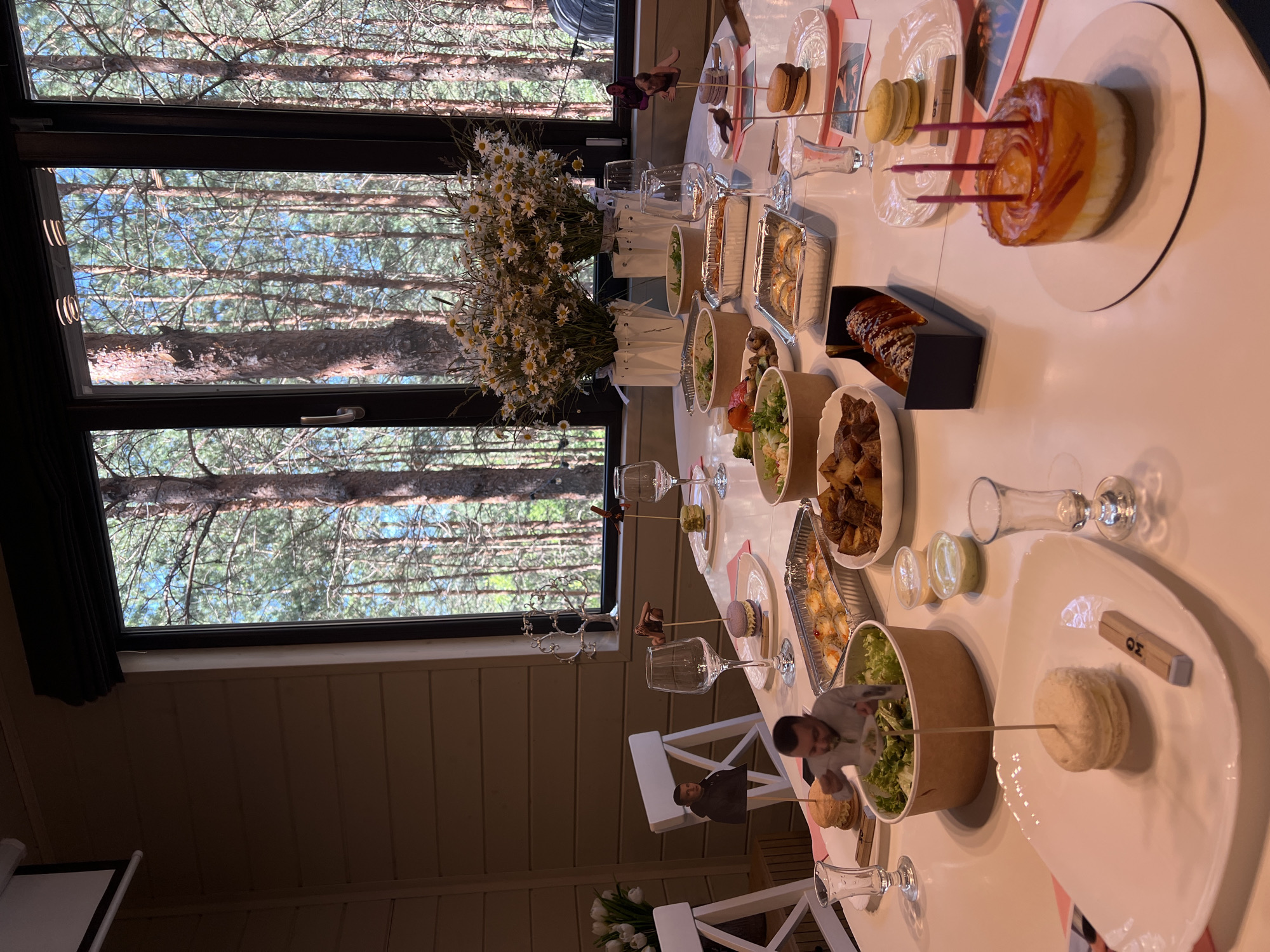 A long, white table is set with food and drinks for a celebration, likely a birthday, as indicated by a cake with candles. The table is positioned in front of a large window that offers a view of a dense forest with tall pine trees. On the table, there are several dishes including a salad in a bowl, a dish of fried potatoes, a container of pasta, a sushi platter, and individual servings of desserts. There are also wine glasses and small shot glasses. Two decorative paper figures of men are placed on skewers, one in front of a burger and another on a chair. A projector screen is visible on the wall above the table. The overall ambiance suggests an indoor gathering with a natural backdrop.