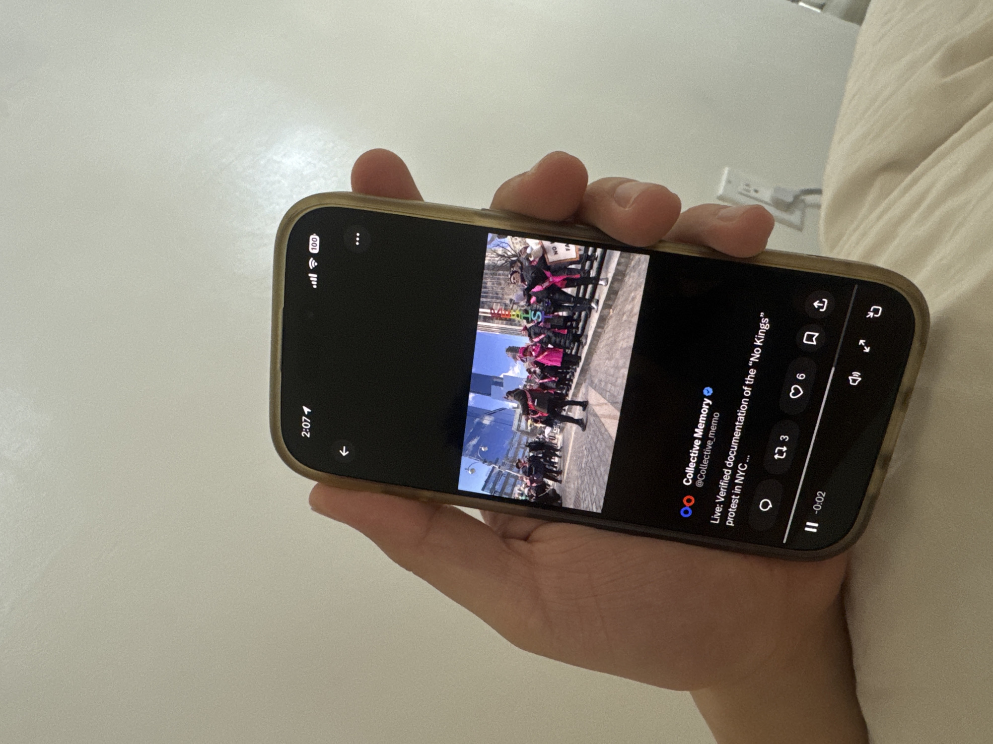 A hand holds a smartphone displaying a social media post. The post is a live video documentation of the "No Kings" protest in New York City, United States. In the video, a group of people can be seen participating in a demonstration. The phone's screen also shows social media engagement metrics. The surrounding environment includes a bed.