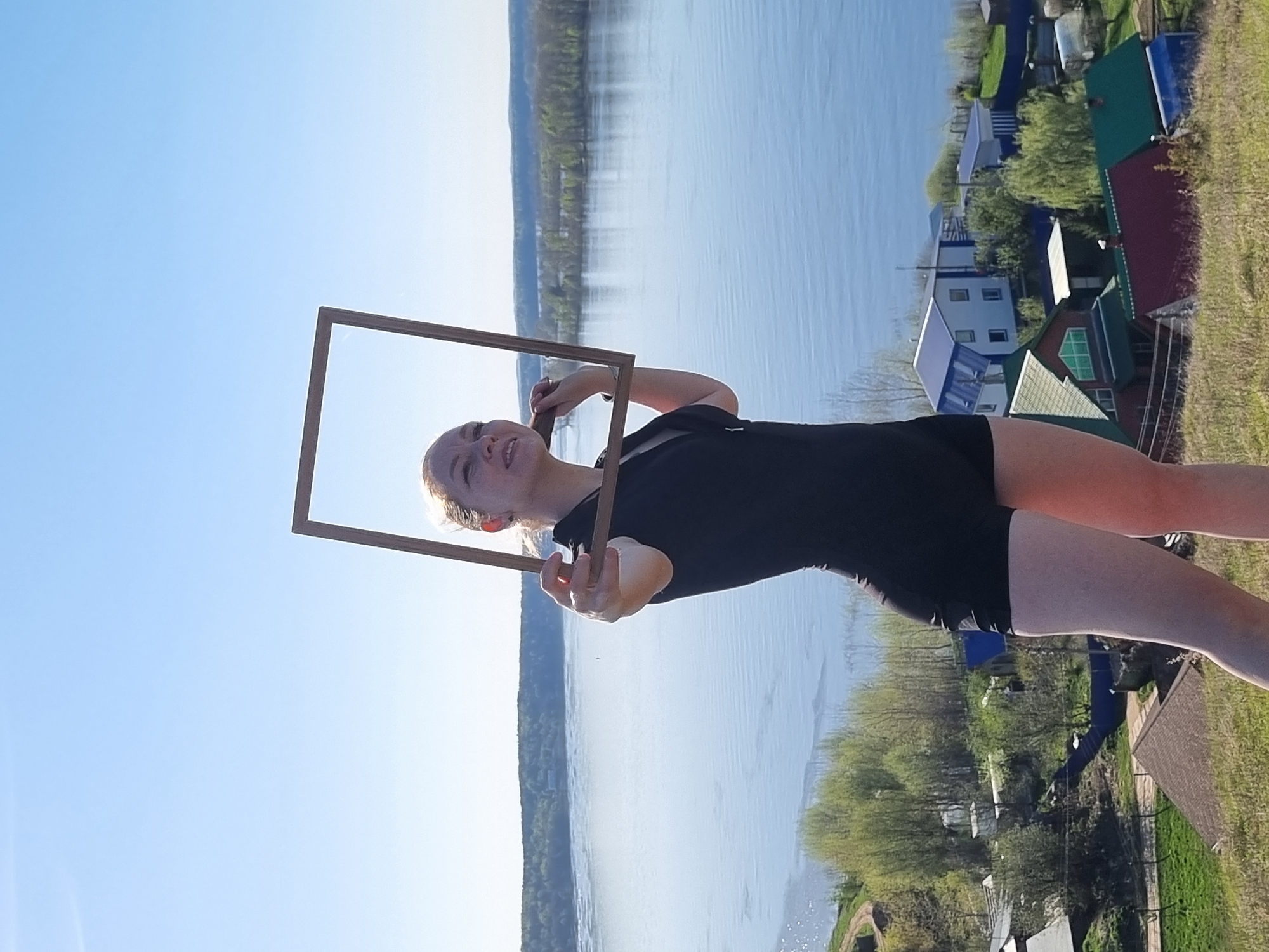 A girl in a black top and shorts stands outdoors in Ольховка, Russia, holding a rectangular wooden frame in front of her. A river extends into the distance behind her and to her left. Houses with colorful roofs are located behind her and to her right. The sky is blue.