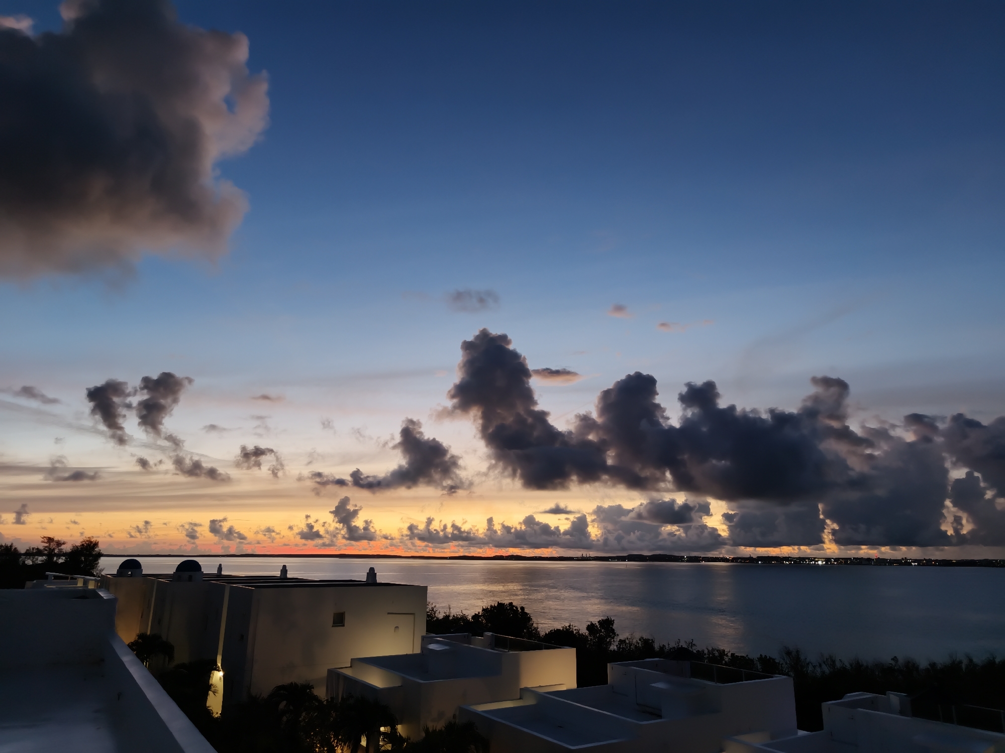 The image captures a twilight scene over a body of water, likely the ocean, with a distant shoreline visible. The sky is a gradient of deep blue at the top, transitioning to shades of orange, yellow, and pink near the horizon, indicative of sunset or sunrise. Dark, dramatic clouds are scattered across the sky, some appearing illuminated by the fading light. In the foreground, the rooftops of several modern, white buildings with sharp architectural lines are visible. These buildings are likely part of a resort or residential complex. The scene is devoid of people. The environment suggests a coastal location with tropical or subtropical vegetation, as indicated by the silhouettes of palm trees. The overall impression is one of tranquility and natural beauty, enhanced by the dramatic sky. The location is identified as Miyakojima, Japan.