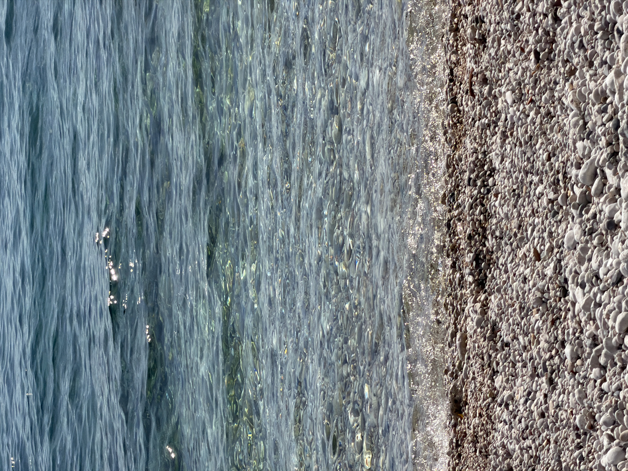 The image displays a serene aerial or overhead view of a clear, calm body of water meeting a pebble beach. The scene is dominated by the natural textures of the water and the shore.
The left side of the image features transparent water, revealing the seabed beneath in various shades of blue and green. The water surface is covered in gentle ripples, creating elongated patterns and reflections of sunlight that shimmer brightly, particularly closer to the shore. The clarity of the water suggests a shallow, undisturbed environment.
To the right, a pristine pebble beach stretches, composed of numerous smooth, rounded stones varying in size from small grains to larger, thumb-sized rocks. The pebbles exhibit a natural palette of grays, whites, and light brown hues. The demarcation between the water and the beach is distinct, with small splashes of foamy water visible along the immediate edge of the shore, likely from gentle waves.
The strong, direct sunlight indicates that it is daytime, possibly morning or early afternoon, contributing to the bright reflections on the water and the well-lit appearance of the pebbles. There are no people, animals, or man-made objects visible, emphasizing the natural beauty and tranquility of the scene. No text is visible in the image. This idyllic coastal setting is located in Grad Rijeka, Croatia.