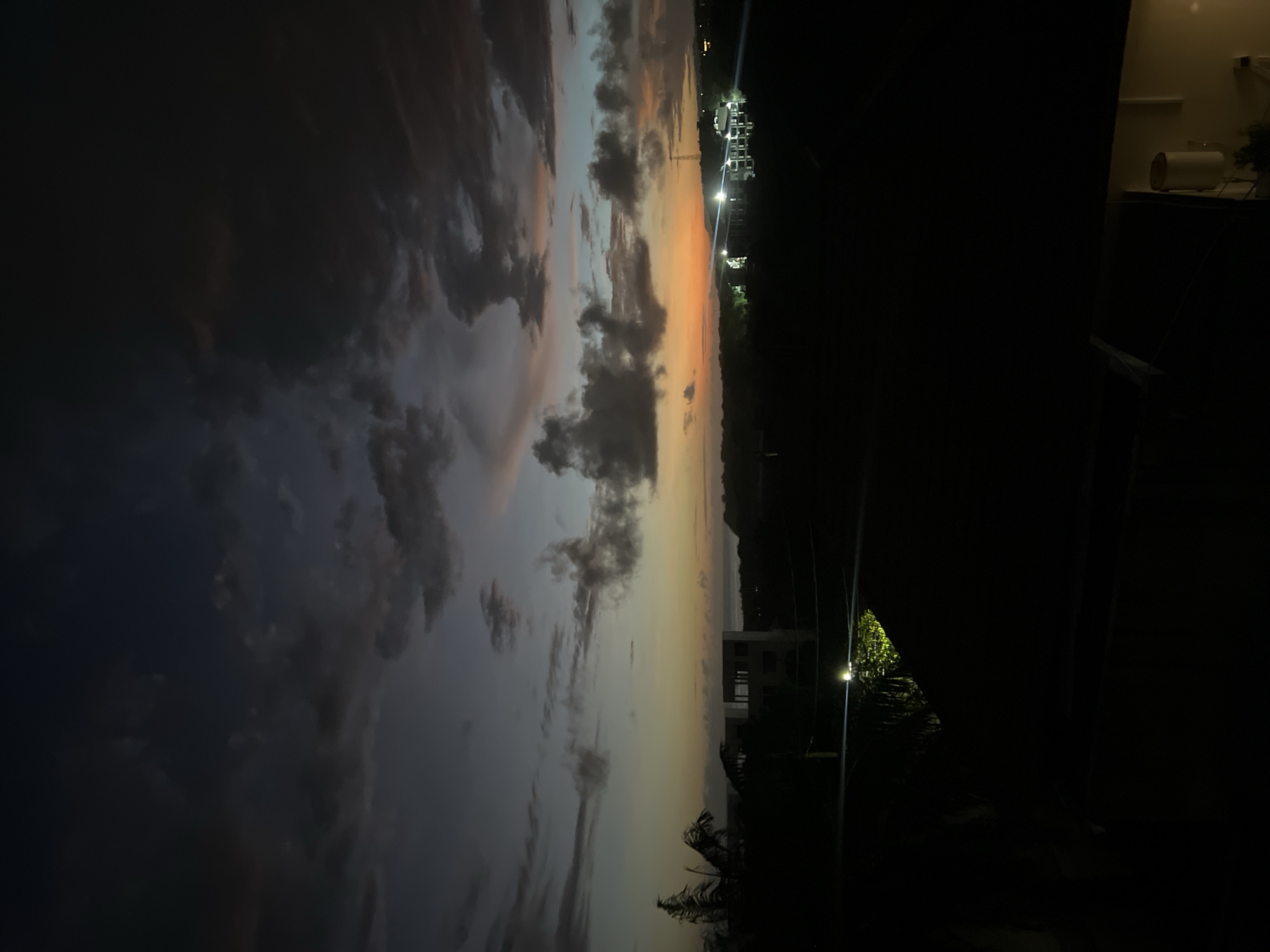 The image depicts a sky at dusk or dawn. The left side of the image shows a sky filled with clouds, some dark and heavy, others lighter with hints of orange. The right side of the image is dark, transitioning into a silhouette of trees, foliage, and a structure with lights in the distance. A light pole is visible in the dark area. The image is taken from what appears to be inside a building, with part of an interior visible in the upper right corner. The location is Unknown, Unknown.
