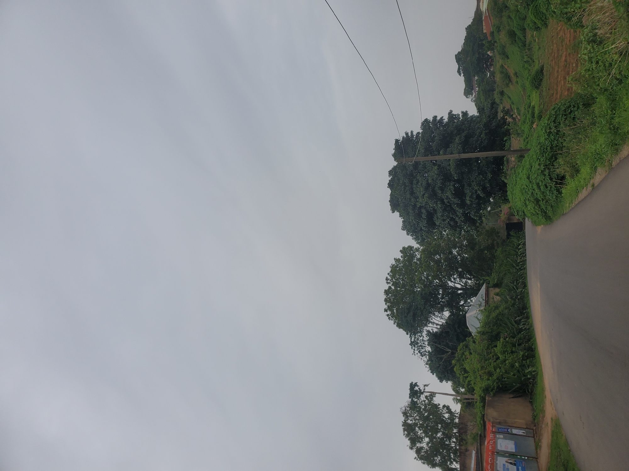 The image depicts a rural road in Jos, Nigeria, under an overcast sky. The road curves to the right, bordered by lush green vegetation and several trees. On the left side of the road, there's a low wall with some posters attached, suggesting proximity to a populated area. Beyond the wall, structures with corrugated roofs and dense foliage are visible. A utility pole with wires stands prominently on the right side of the road, surrounded by thick bushes and a gently sloping, uncultivated field. The overall scene conveys a sense of quiet, everyday life in a semi-urban or rural setting.