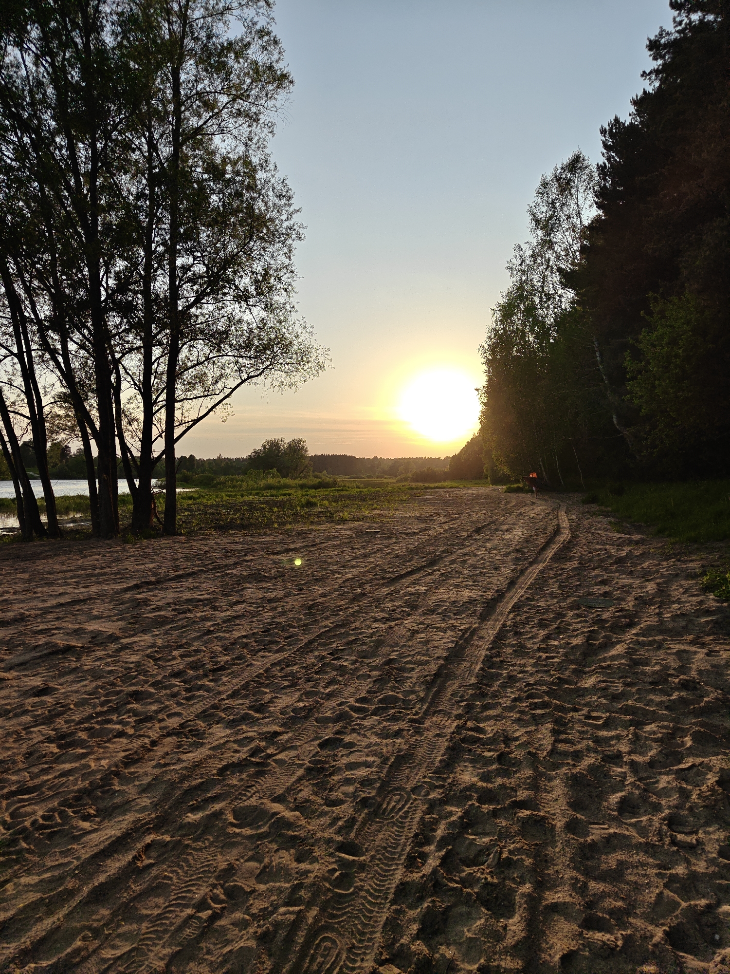 The photograph captures a sandy beach at sunset in Мышкино, Russia. The sky is a gradient of light blue to orange, with the sun prominently positioned near the horizon. To the left, a stand of slender trees casts shadows on the sand. A body of water is visible behind the trees, suggesting a river or lake. To the right, denser forest borders the beach. Tire tracks run through the sand, leading towards the setting sun. The sand itself is textured with numerous footprints and other impressions. In the distance, a small figure in an orange shirt appears to be walking along the path. The overall scene conveys a sense of tranquility and natural beauty.